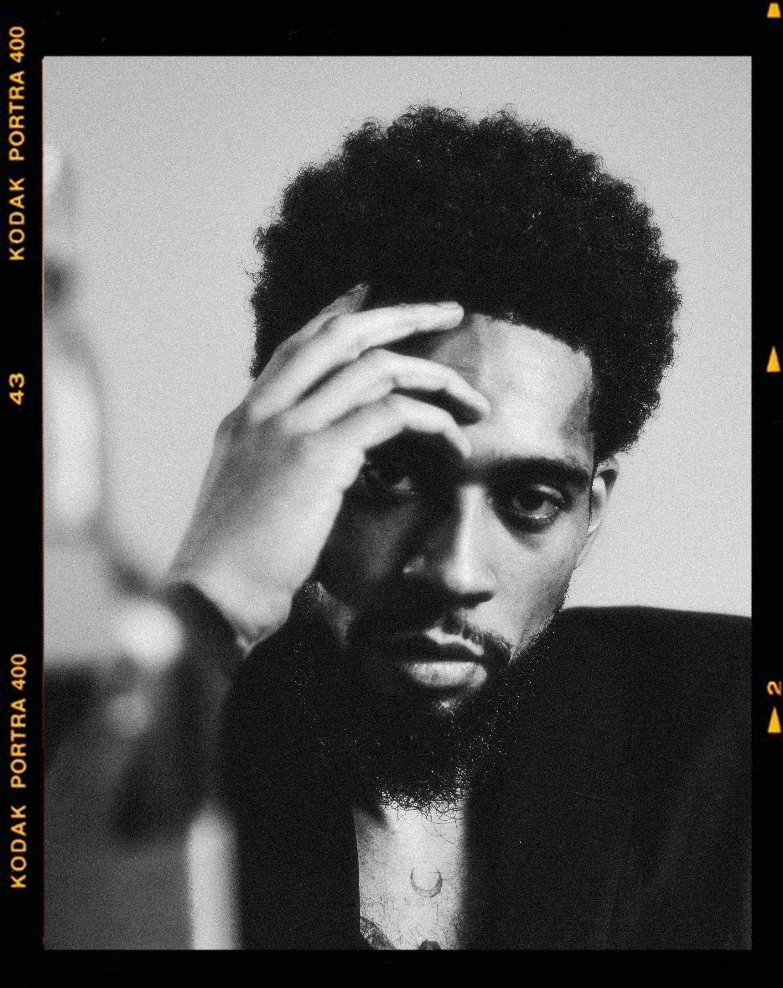 Black and white portrait of a man with curly hair and a beard, holding his hand up to his forehead, looking intensely at the camera.