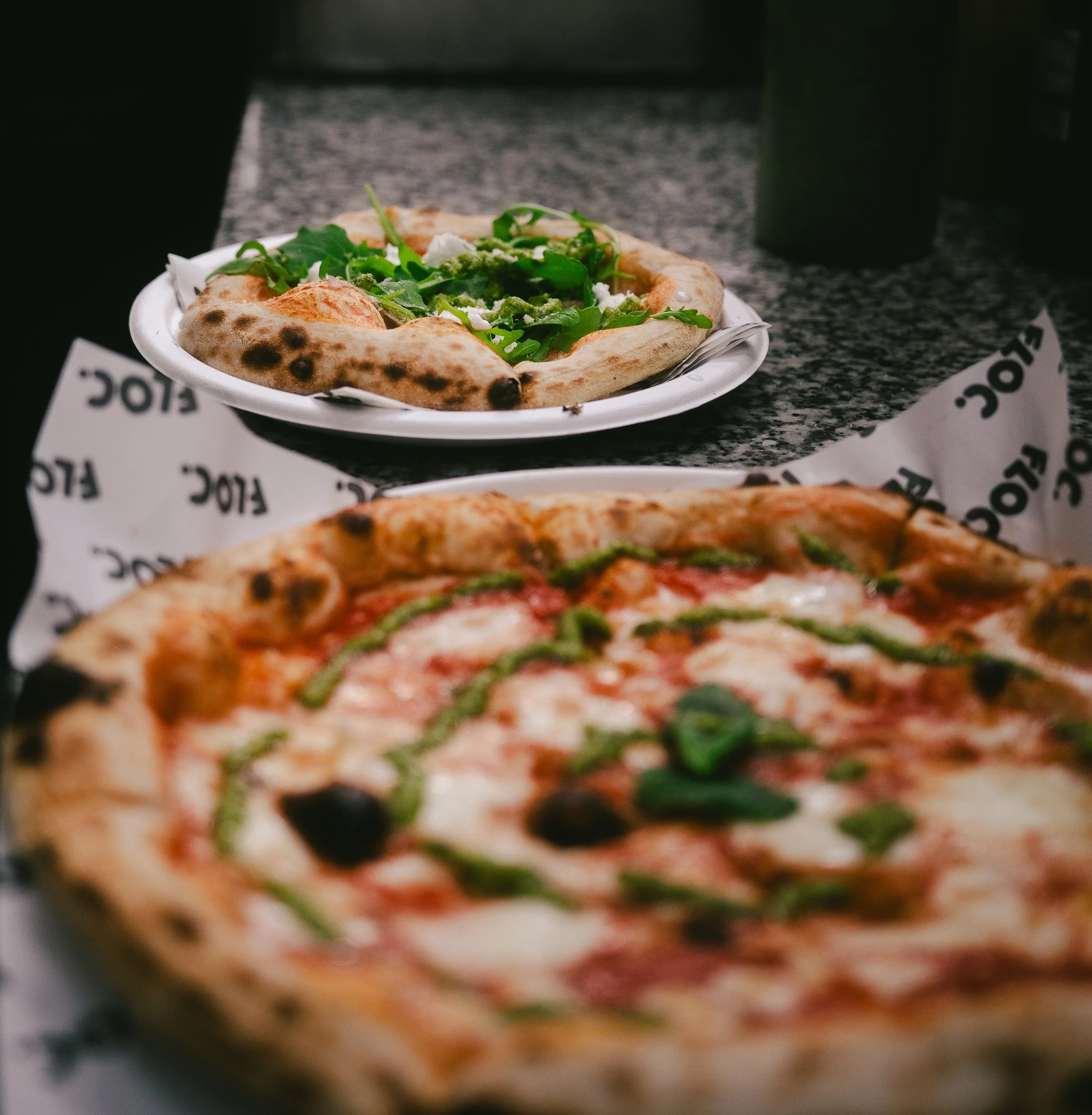 Close-up of a pizza with green peppers, black olives, and cheese in the foreground, and a plate with a pizza and arugula salad on a plate in the background on a dark speckled countertop.