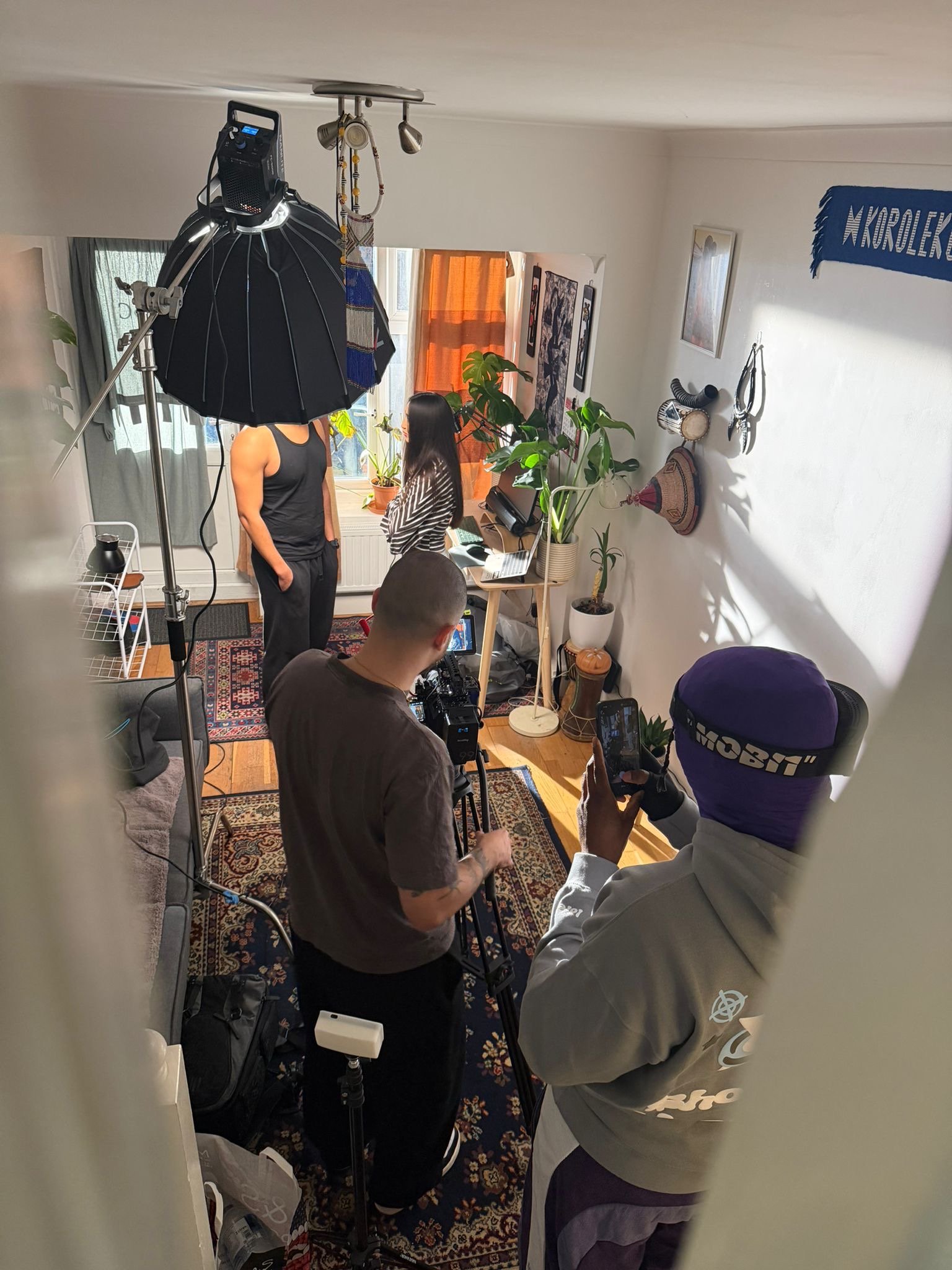 A behind-the-scenes shot of a photo or video shoot inside a small room. Two people are on camera, with one man and one woman standing near a window. Two crew members are operating equipment: one adjusting a camera and another managing lighting or sound. The room is decorated with plants, artwork, and a blue banner on the wall.