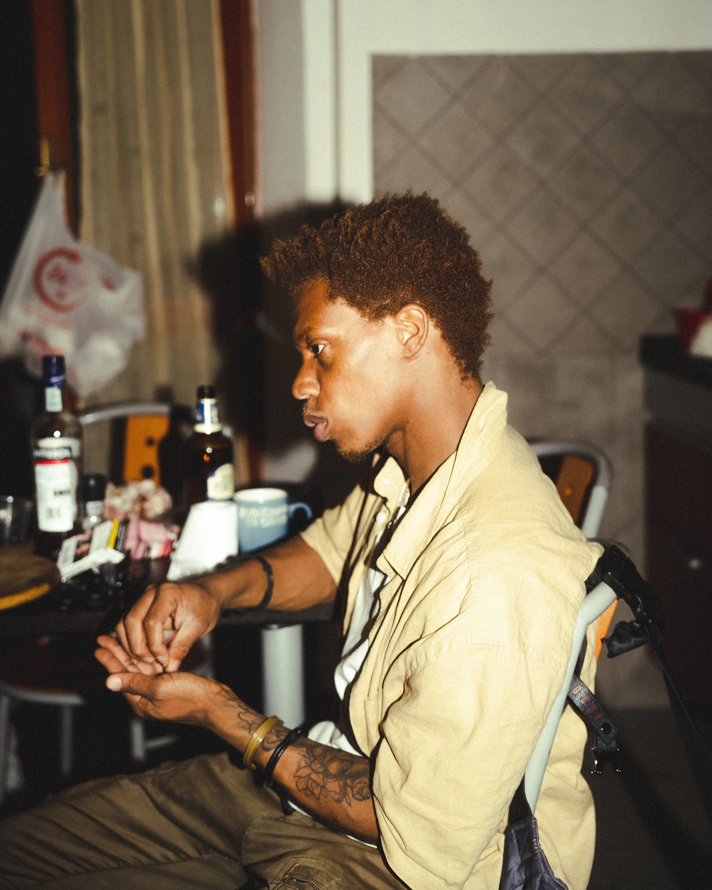 Young man with tattoos sitting at a cluttered table in a cozy room, focused on something in front of him.