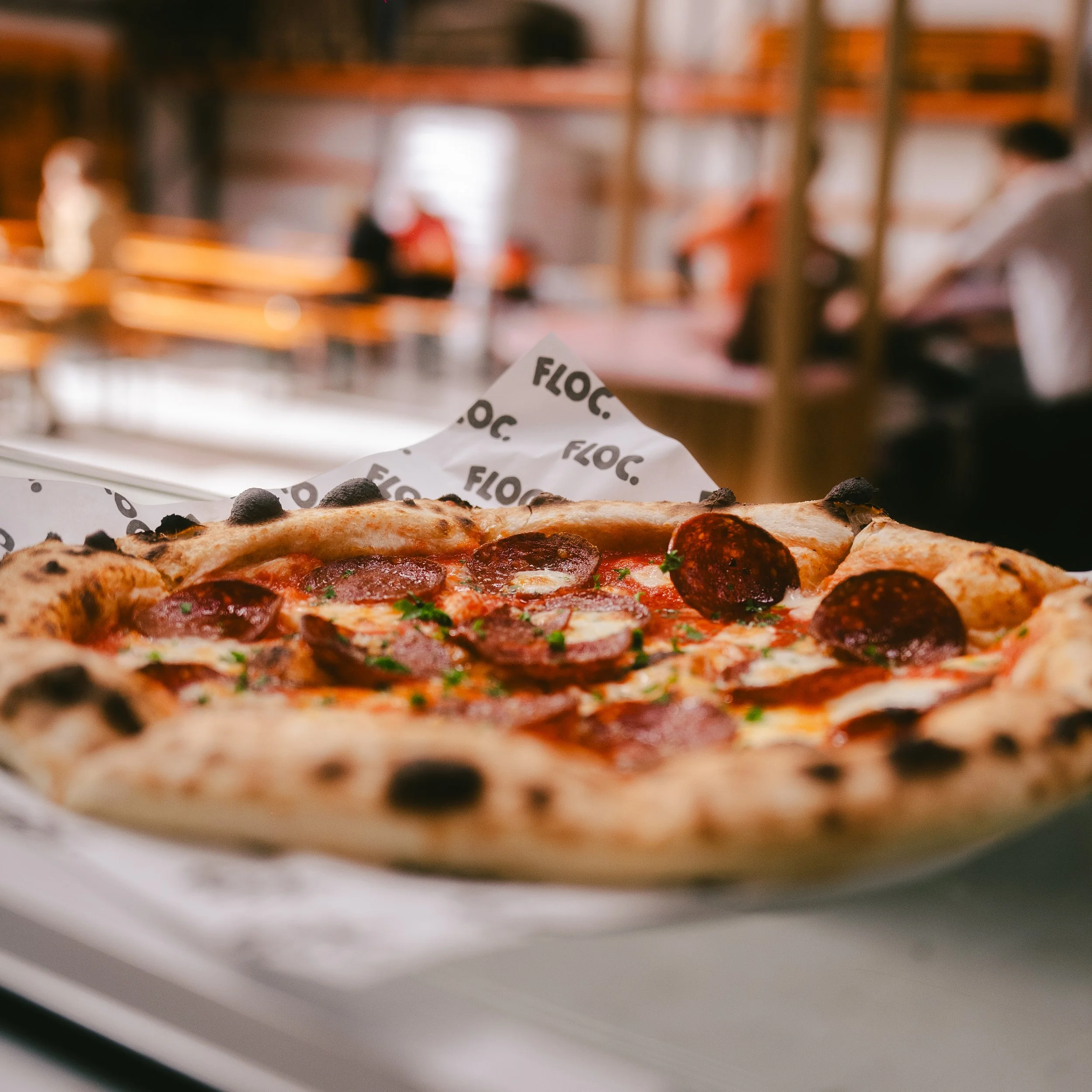 Close-up of a pepperoni pizza on a table in a restaurant with wooden tables and blurred customers in the background.