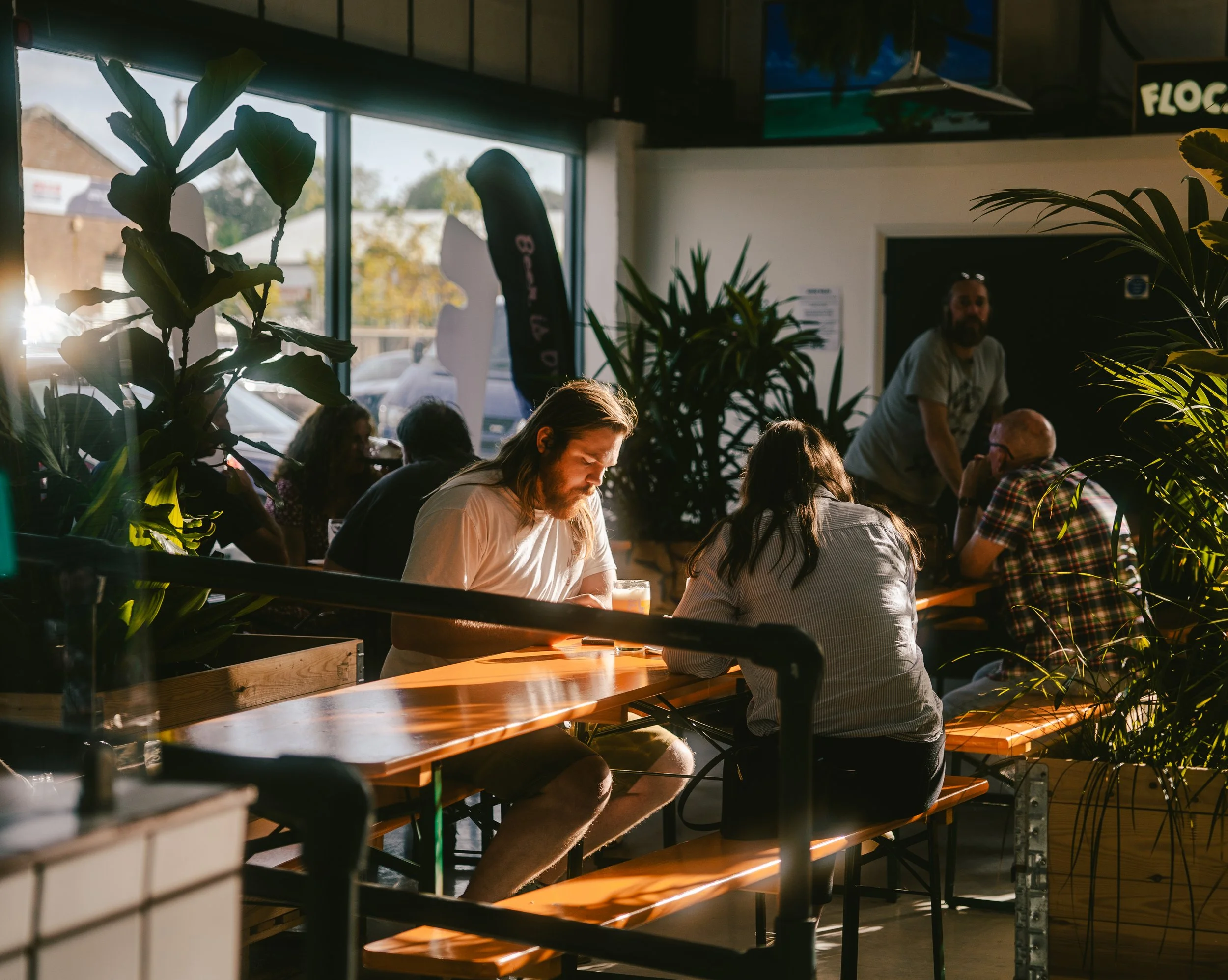 People sitting at a long wooden table inside a cafe, illuminated by sunlight through large windows, with plants and a staff member in the background.