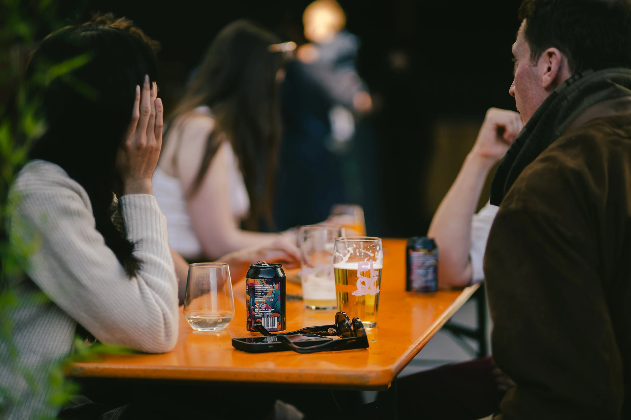 A group of people sitting at a wooden table with drinks, with a woman covering her face and others blurred in the background.