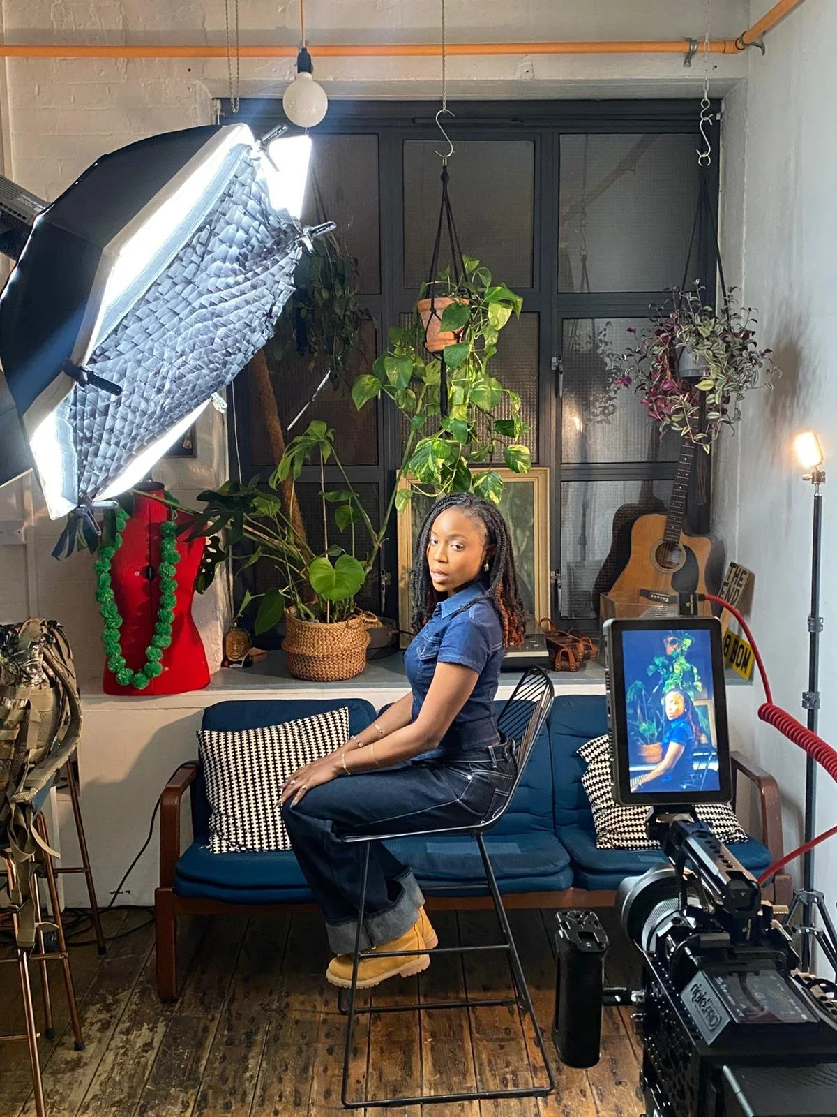 A woman, wearing a denim shirt and jeans, sitting on a barstool in front of a camera setup with studio lights. The background features a black wall, large window, hanging plants, a guitar, a portrait, and decorative pillows on a blue couch.