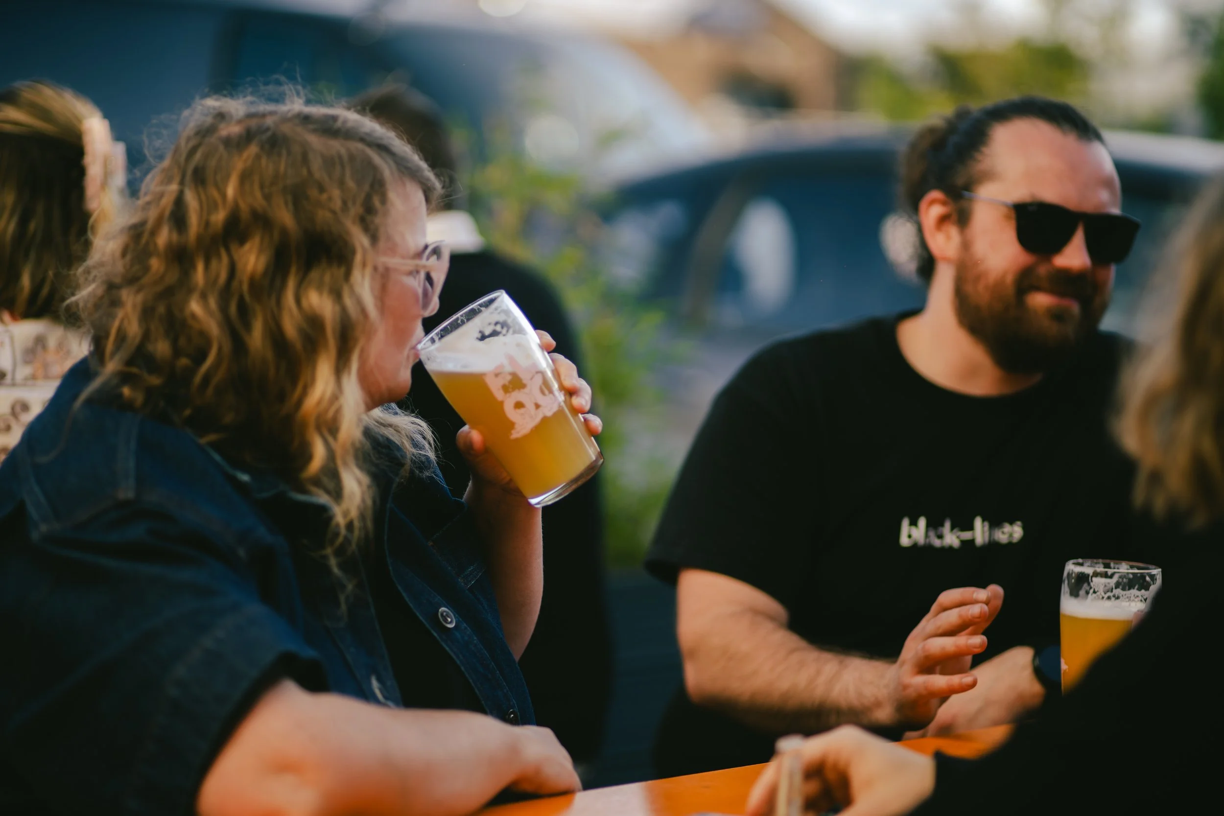 People socializing outdoors at a bar or restaurant, holding glasses of beer, with a woman in the foreground drinking beer and a man wearing sunglasses in the background.