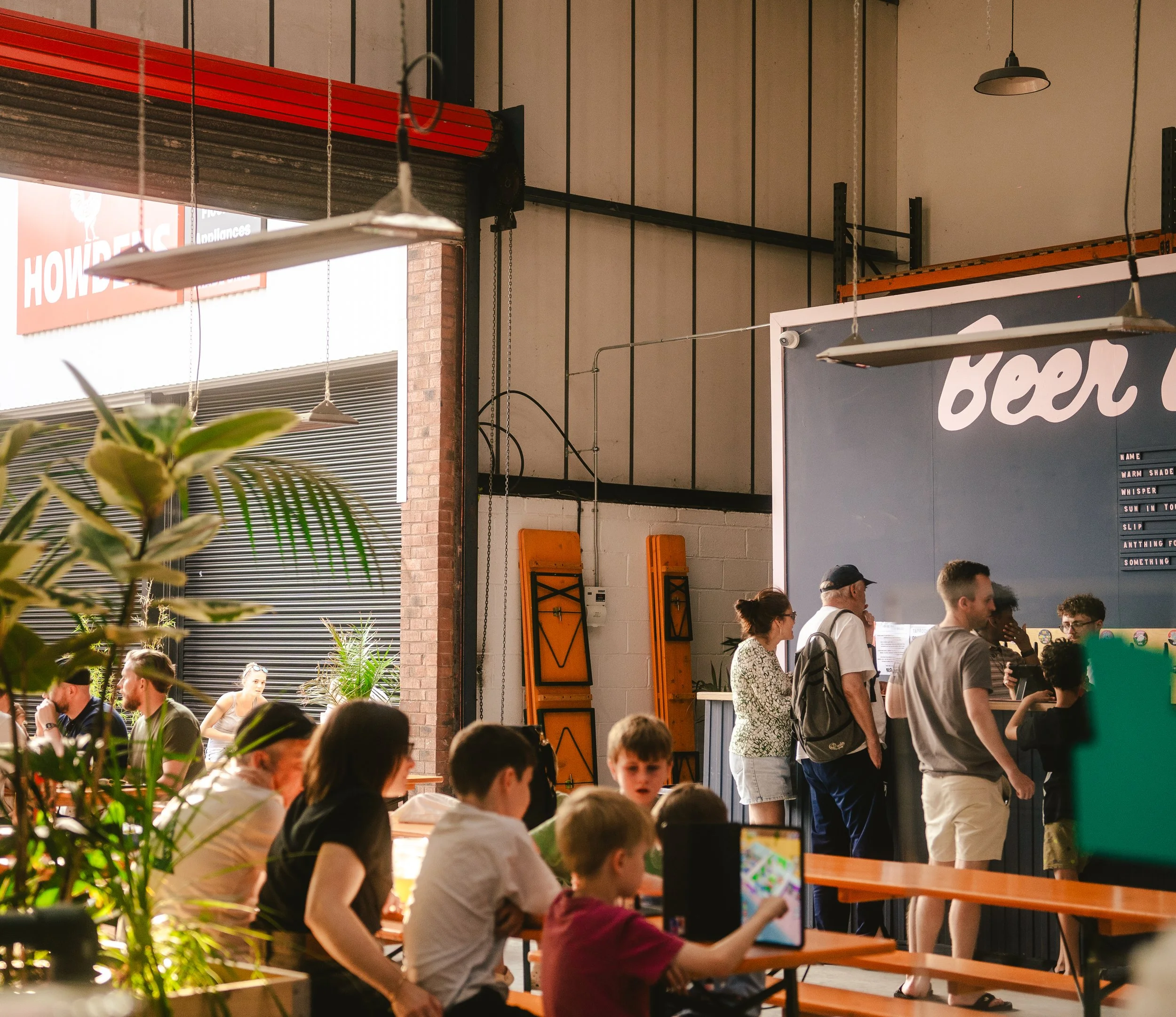 People wait in line and sit at tables inside a restaurant or cafe, with some children playing and others using devices, while sunlight streams in through large open doors.