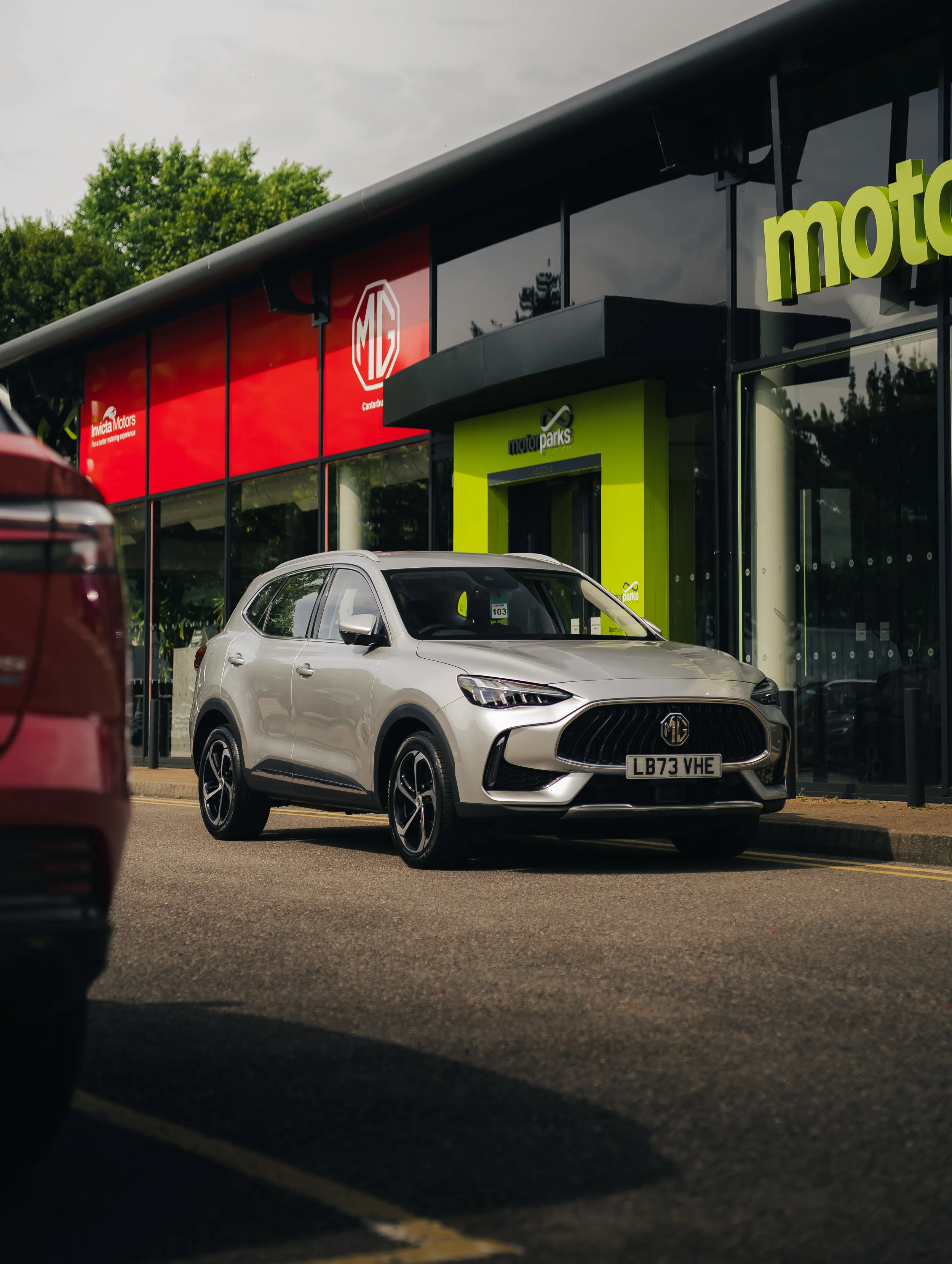Silver MG SUV parked outside a motorsport dealership with MG and motor parks signs, and a red MG sign on the building.