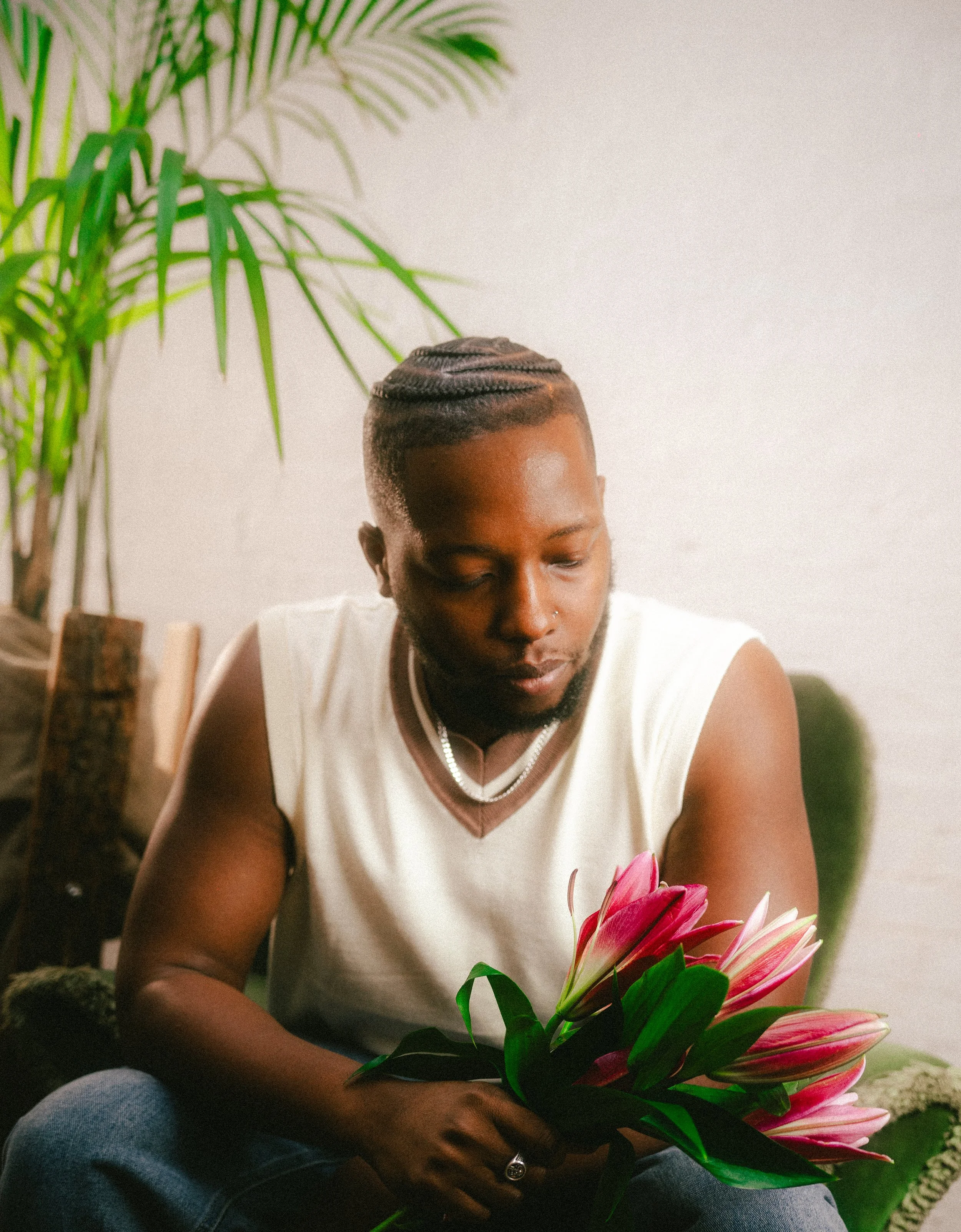 A man with braided hair and a goatee sitting on a green sofa, holding pink lilies, with a large green plant in the background.