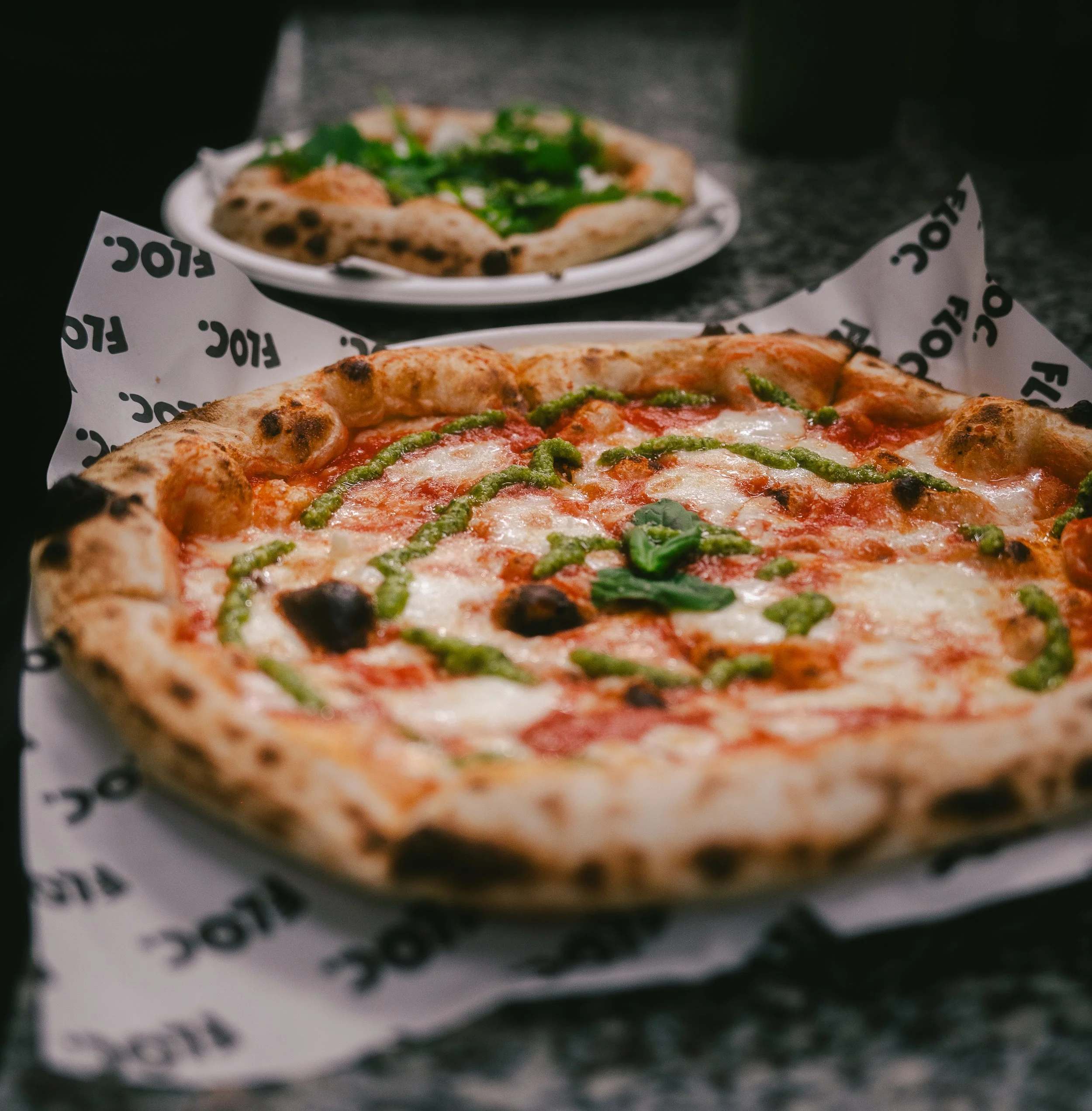 Close-up of a pepperoni pizza with green basil and drizzle of green pesto on top, on a paper-lined tray. In the background, a plate with a smaller pizza with green toppings, possibly asparagus or green peppers, on a dark countertop.