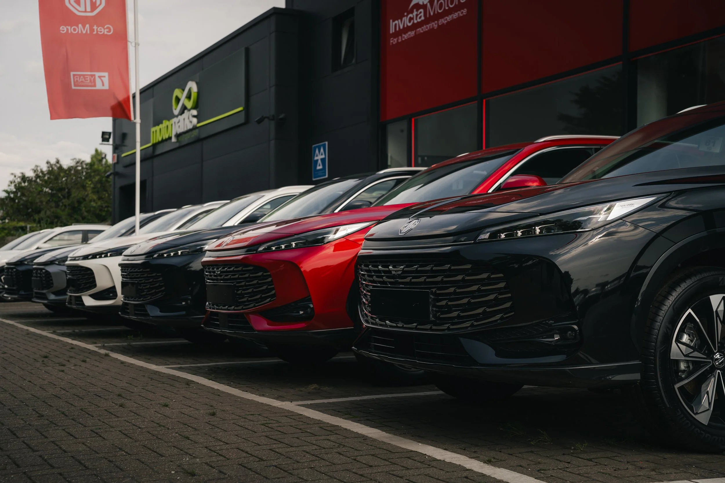 Row of new cars parked in front of a dealership building with a large dealership logo and signage, including a red banner and red and black building exterior.