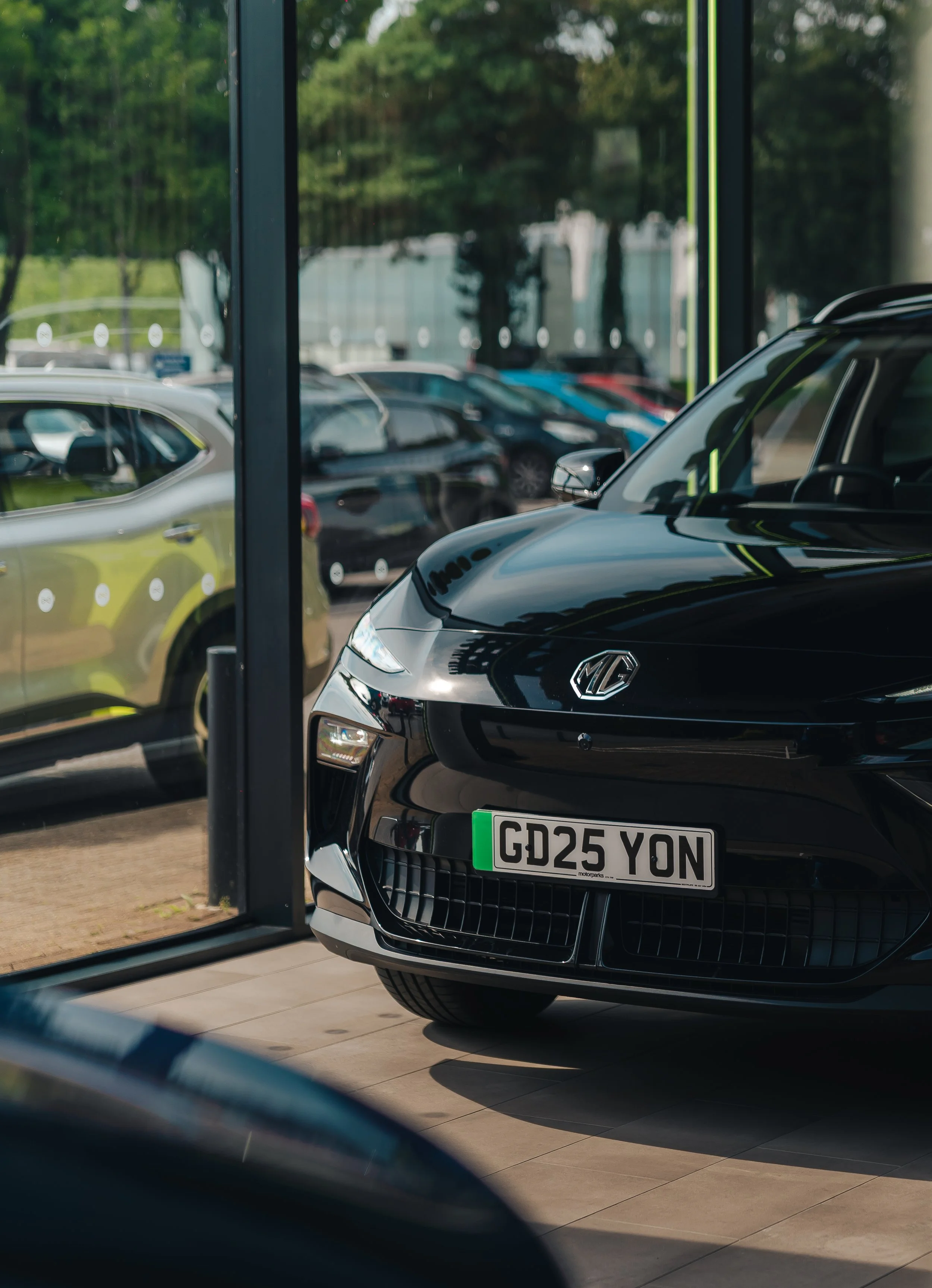 Black MG car with a UK registration plate reading 'GD25 YON' inside a showroom, reflected on the glass window, with cars and trees visible outside.
