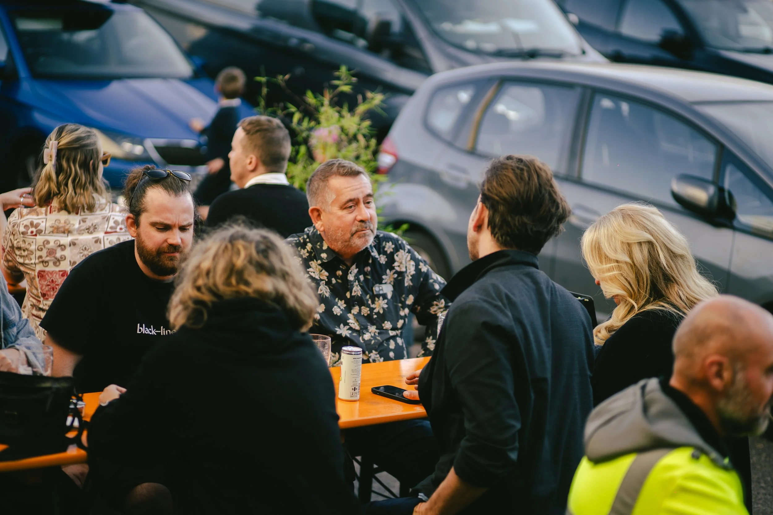 People sitting at a table outdoors, engaging in conversation, with parked cars in the background.