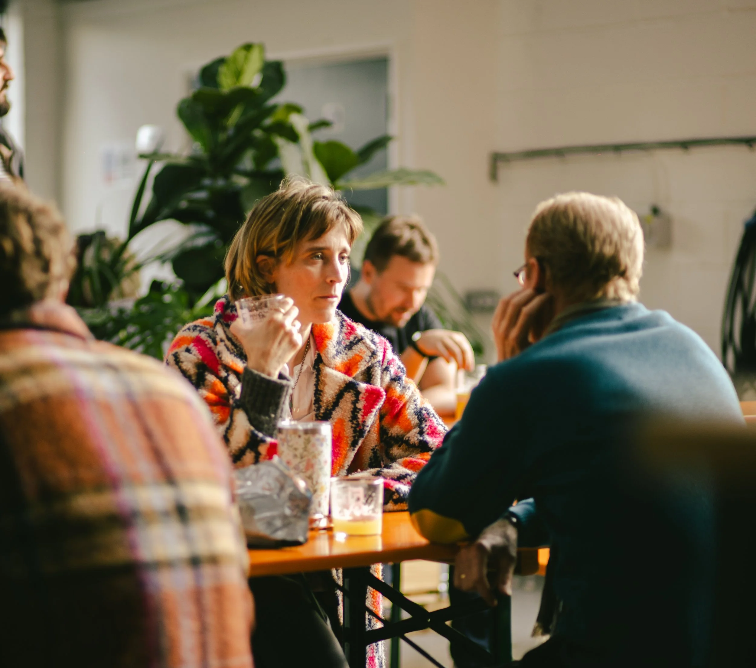 A group of people sitting around a table having a conversation in a cozy, well-lit indoor space with large green plants in the background.