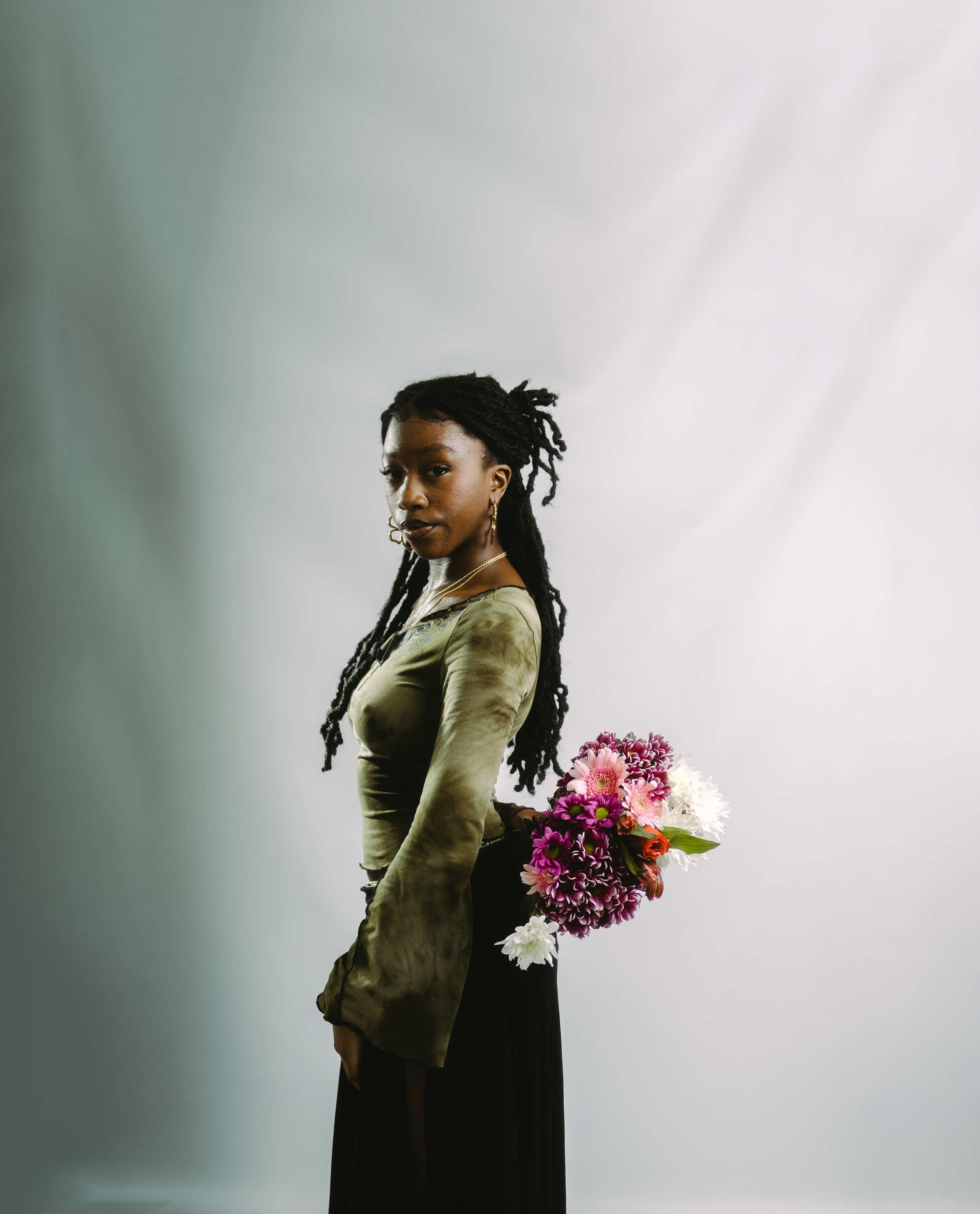 A woman with long dreadlocks stands sideways, holding a colorful bouquet of flowers behind her back, wearing a long-sleeved top and a black skirt, against a gray background.