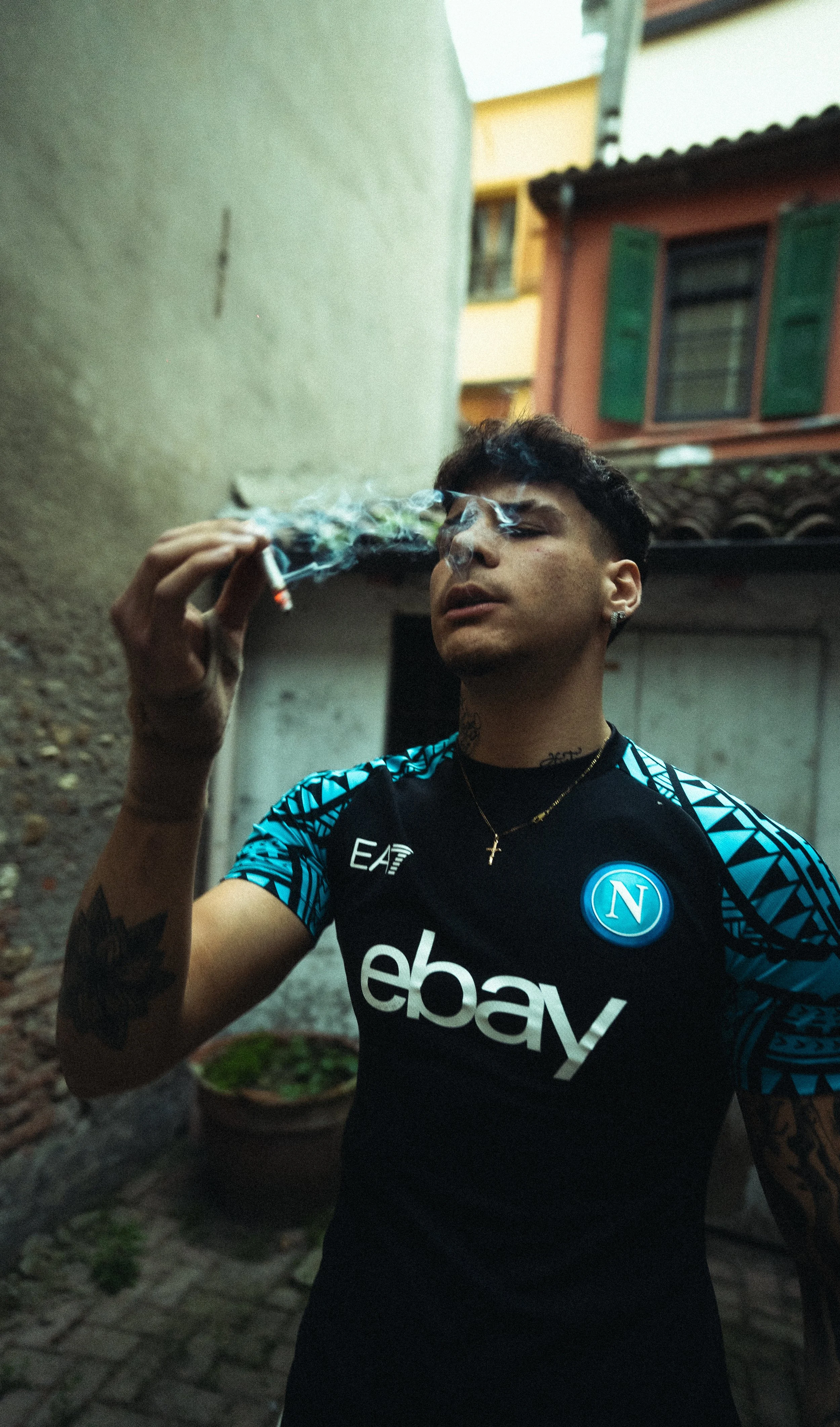 Young man with tattoos on his arm and neck, wearing a Napoli football jersey, exhales cigarette smoke in an alleyway with colorful buildings in the background.
