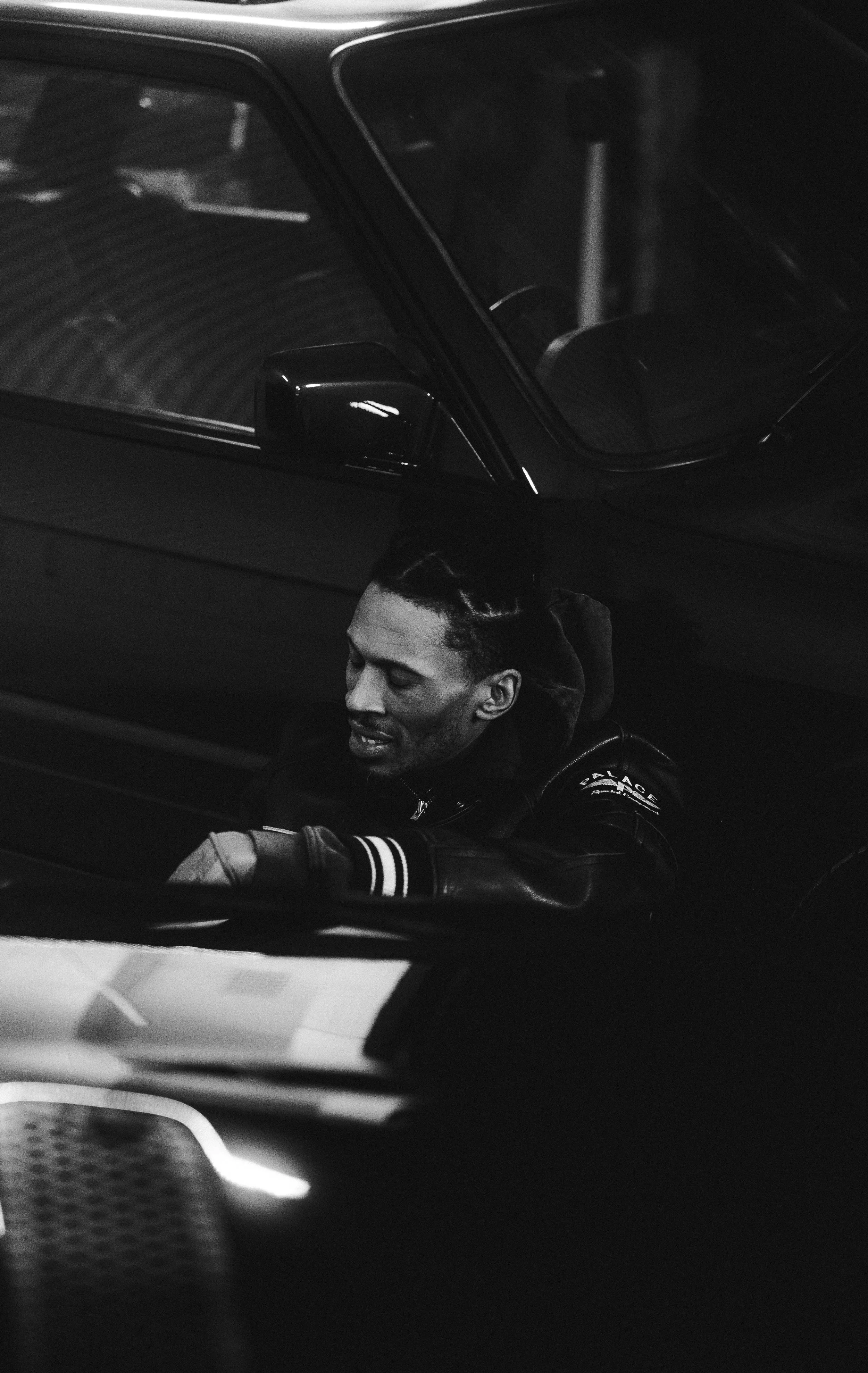 Black and white photo of a man with braided hair sitting in a car, looking down, with the car door slightly open.