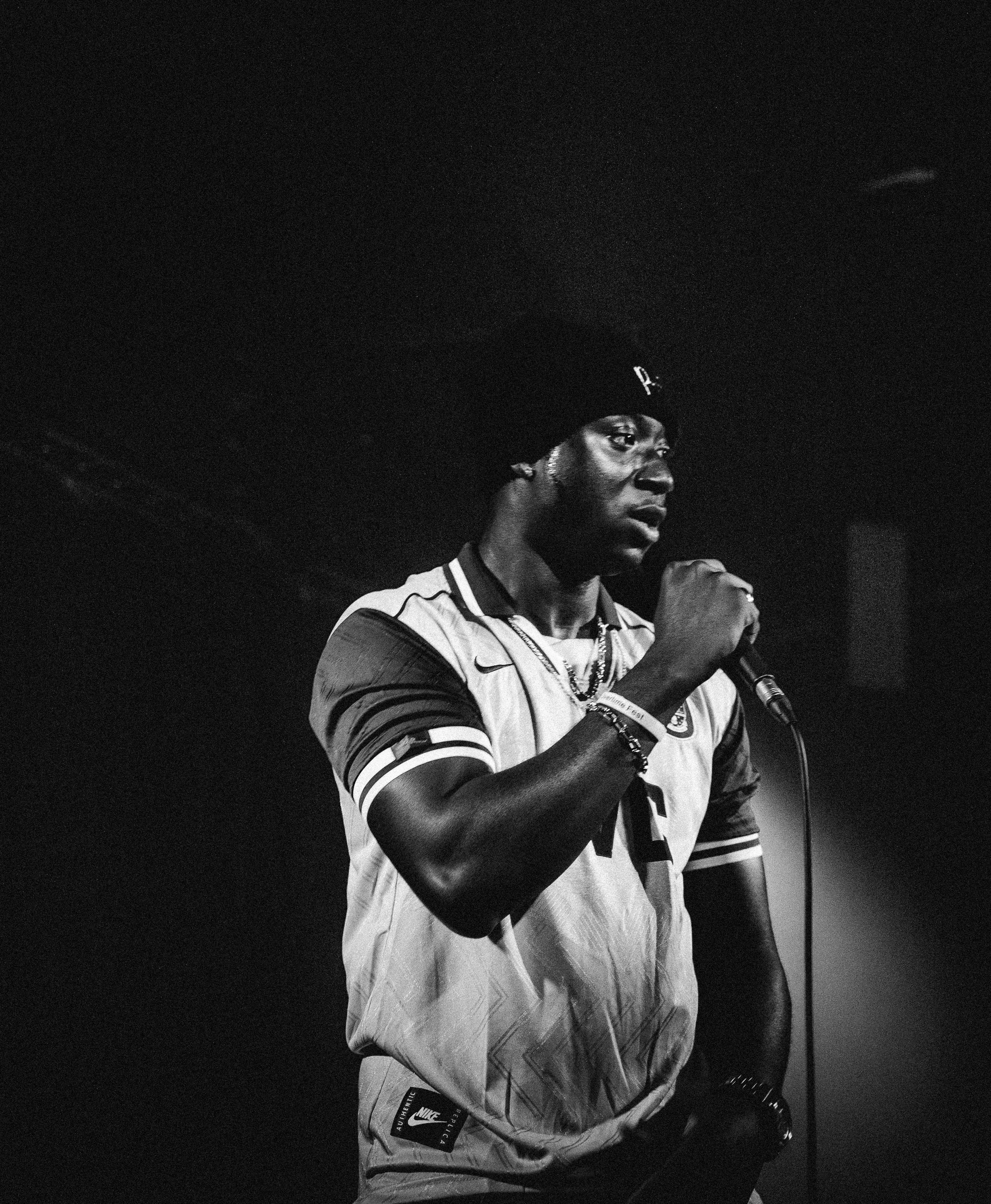 A Black man wearing a Nike sports jersey, beanie, and jewelry, holding a microphone, standing in a dark environment.