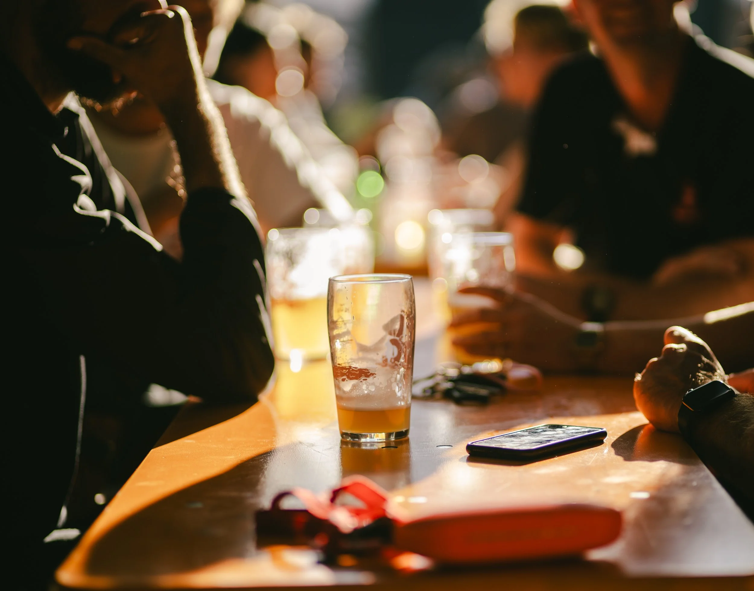 People sitting at a wooden table outdoors during daytime, with glasses of beer, a smartphone, and a red face mask on the table.