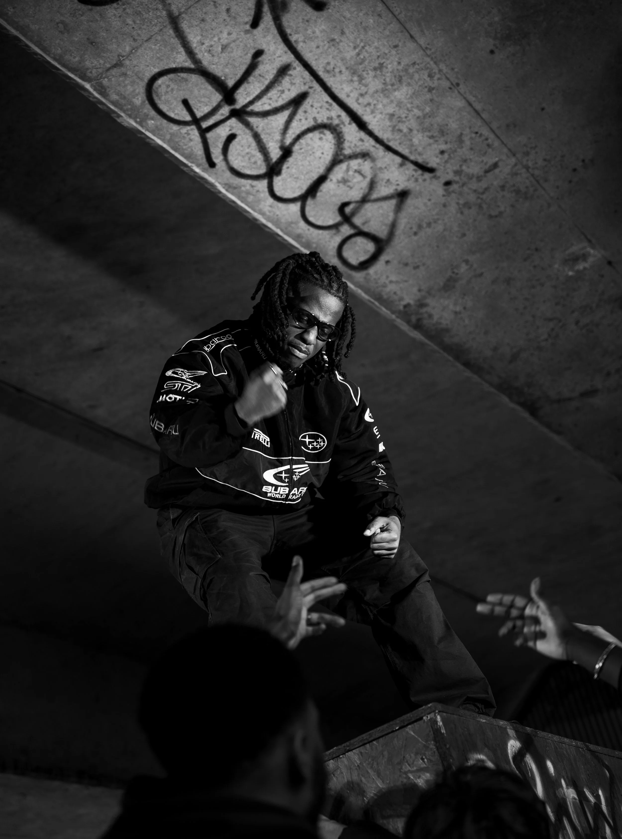 A man with dreadlocks and sunglasses, wearing a jacket with racing logos, is performing a dance move on a platform, with a graffiti-covered ceiling above him, in an urban setting.