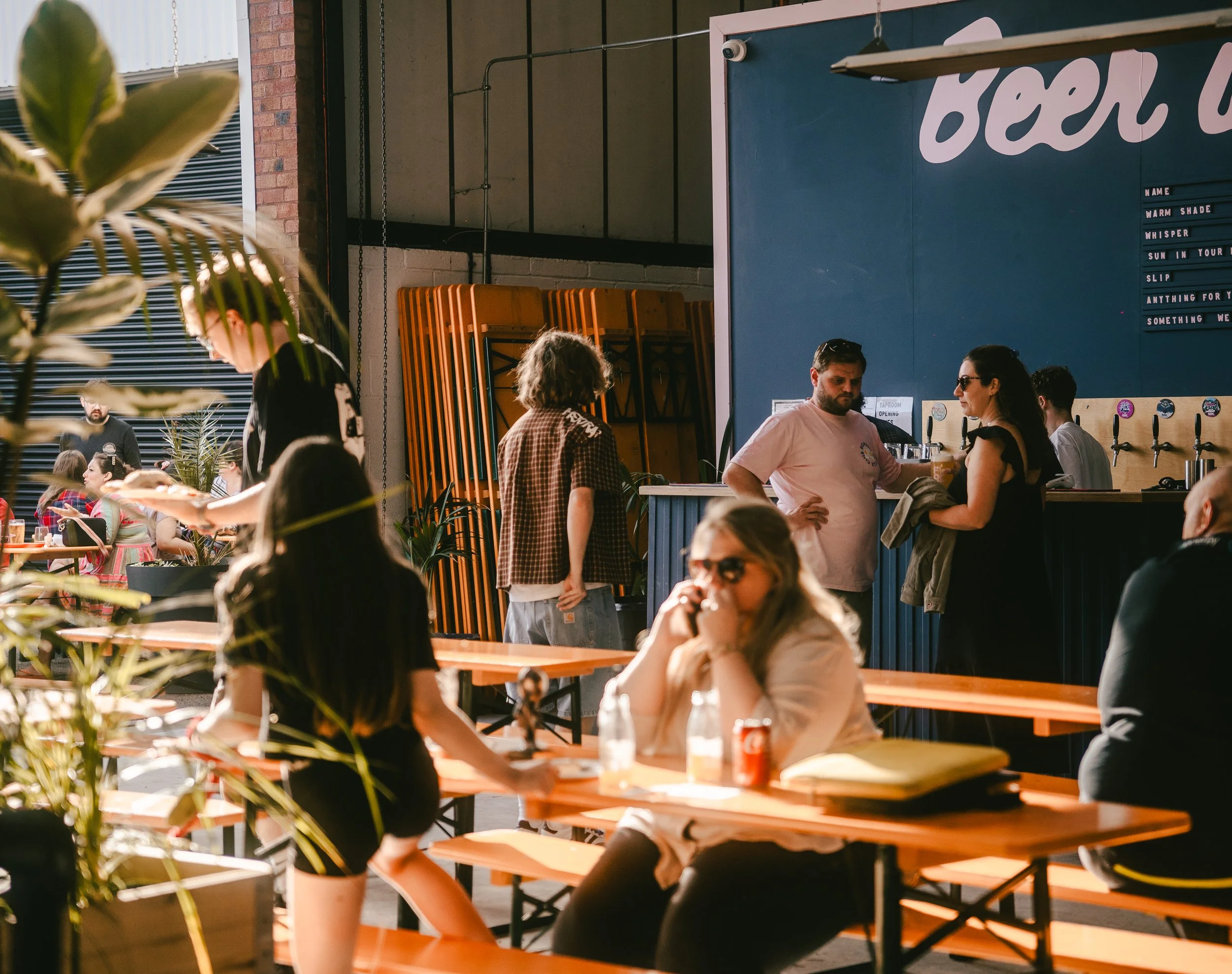 People dining and standing at a bar inside a modern, brightly lit establishment with orange tables and chairs, plants, and a blue wall with decor.