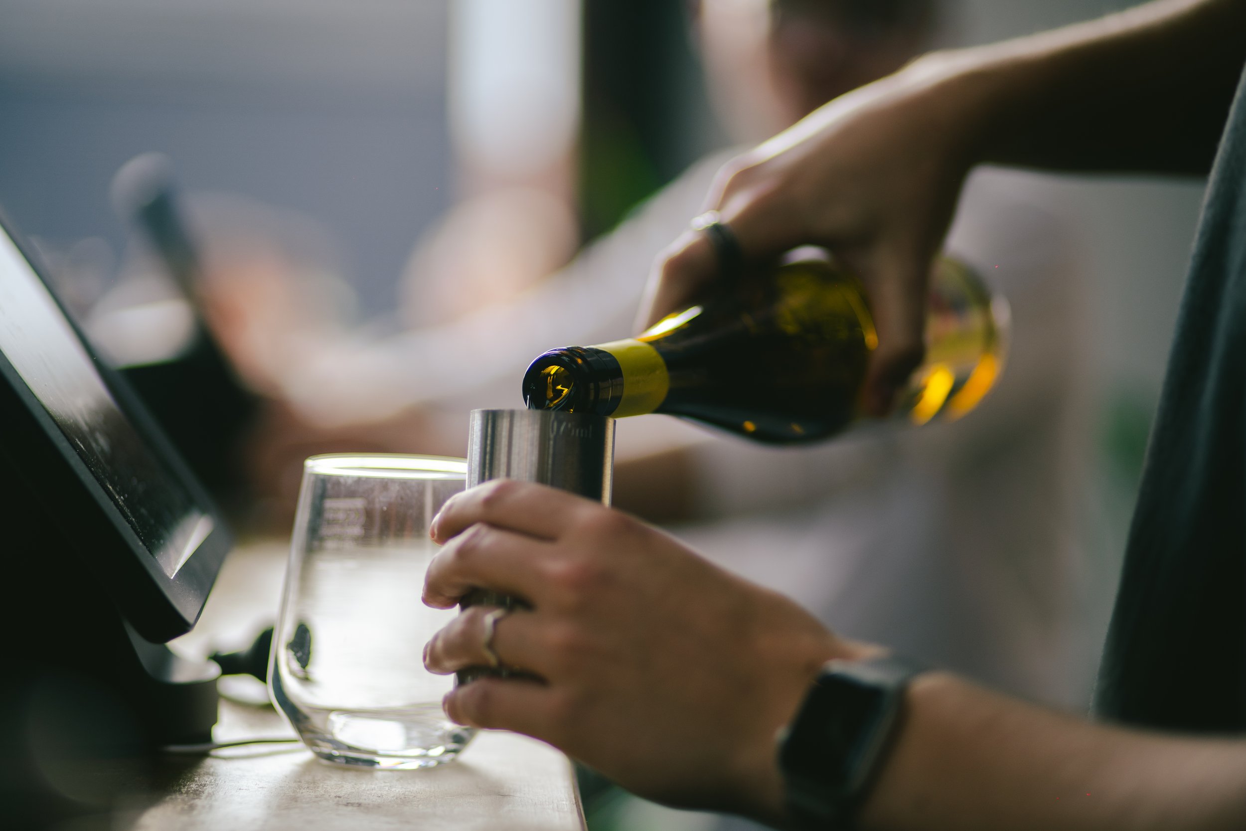 Person pouring beer from a bottle into a glass on a counter near a computer monitor.