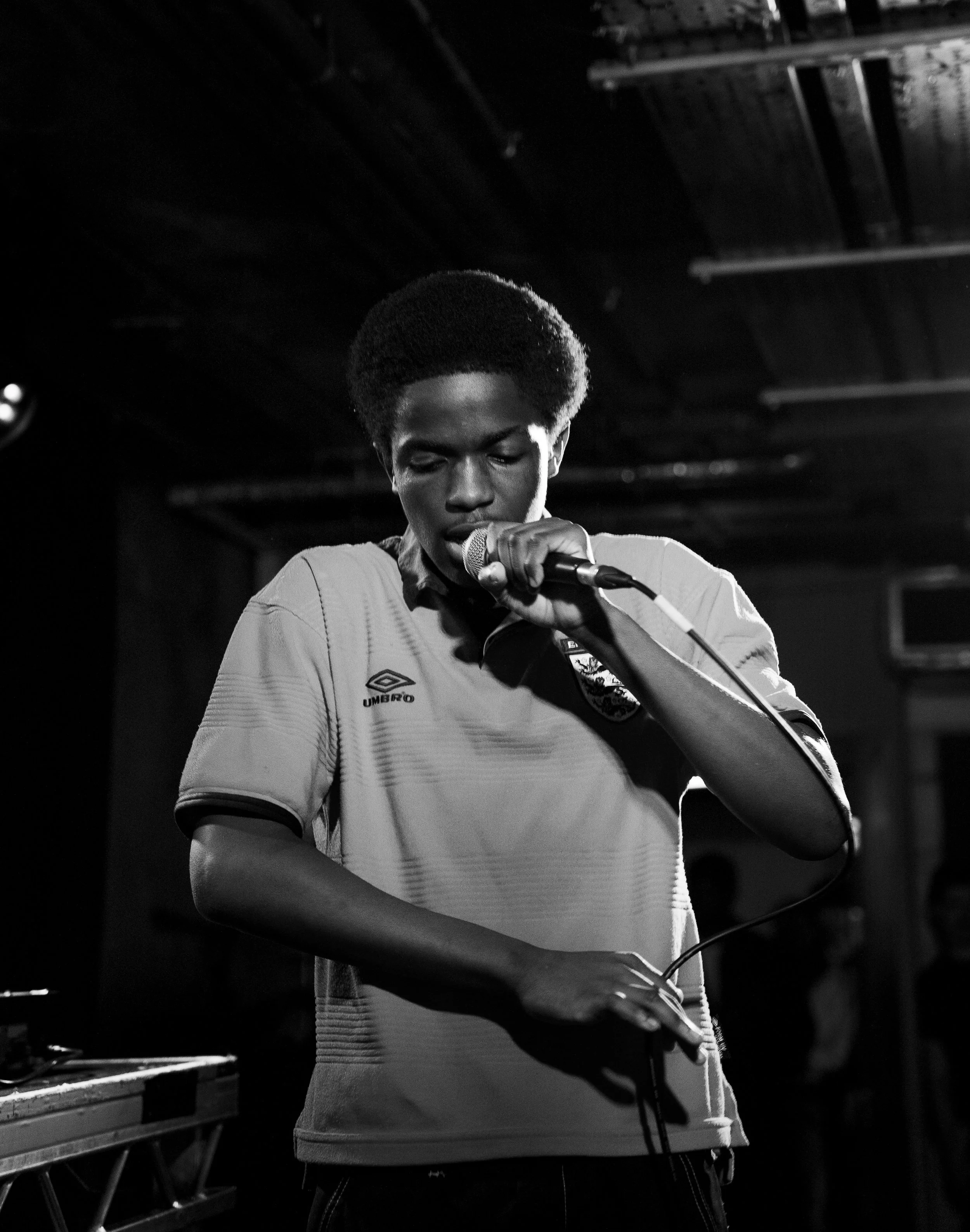 Black and white photo of a young man with an afro hairstyle holding a microphone and a small electronic device, possibly a smartphone, in a dark indoor setting with a low ceiling and some equipment in the background.