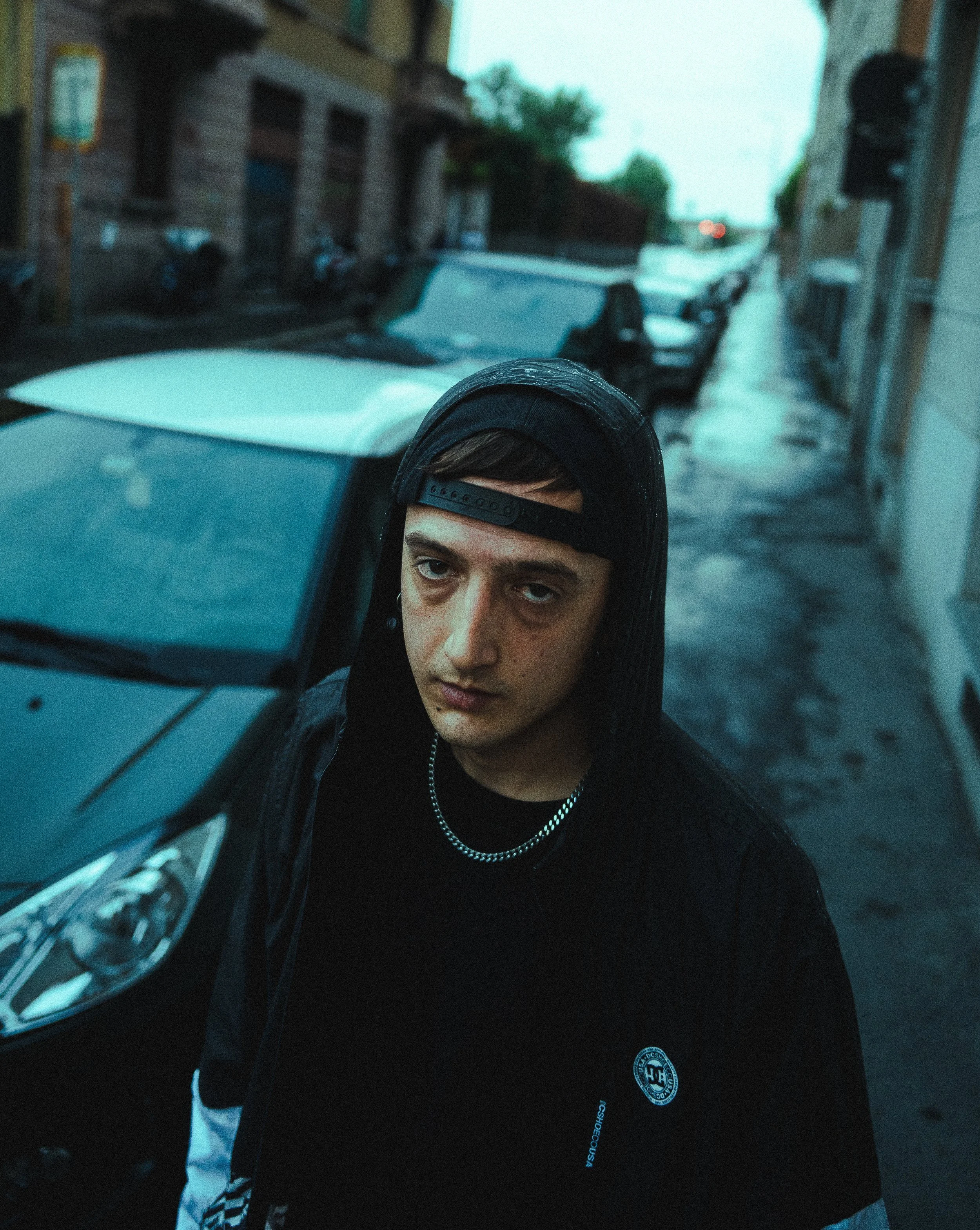 A young man with dark hair and freckles looking directly at the camera, wearing a black cap backwards, a chain necklace, and a black hoodie with a small logo, standing on a wet city street with parked cars and buildings.