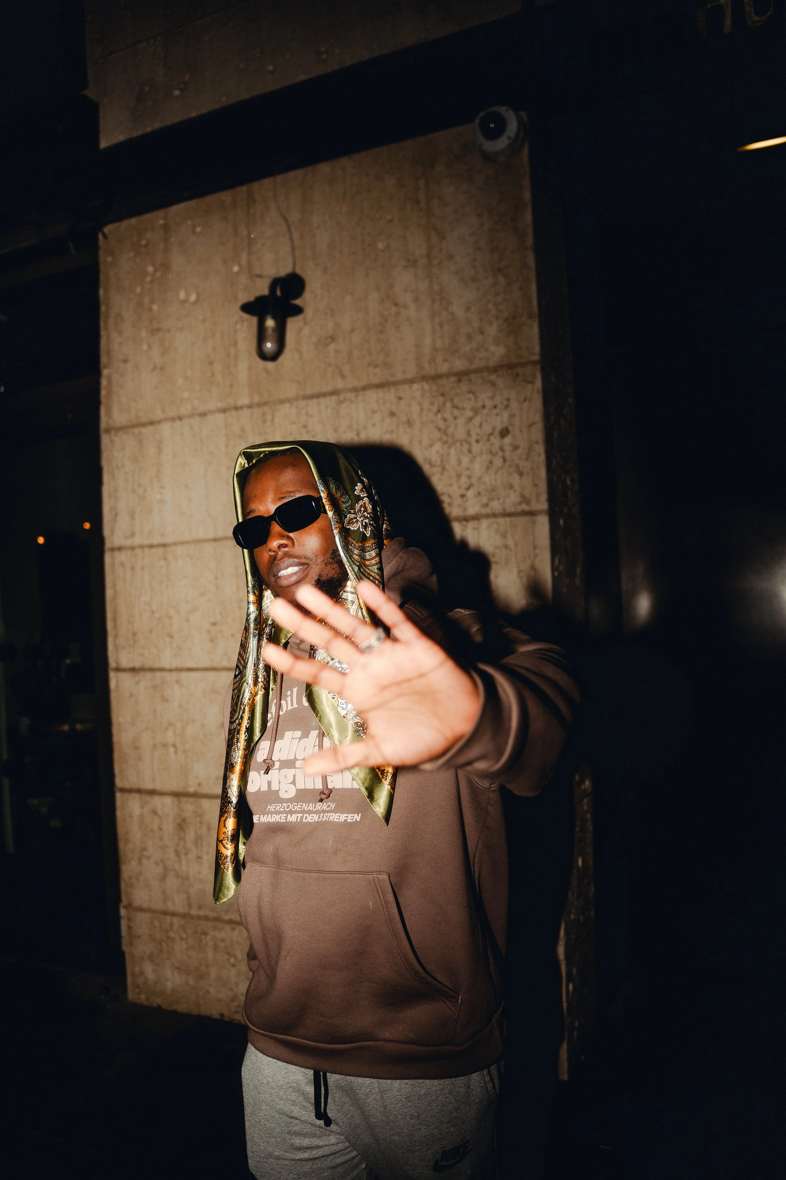 A man wearing sunglasses and a scarf around his head, dressed in a brown hoodie and gray sweatpants, standing outdoors at night. He is holding up his hand as if to stop or signal to someone.