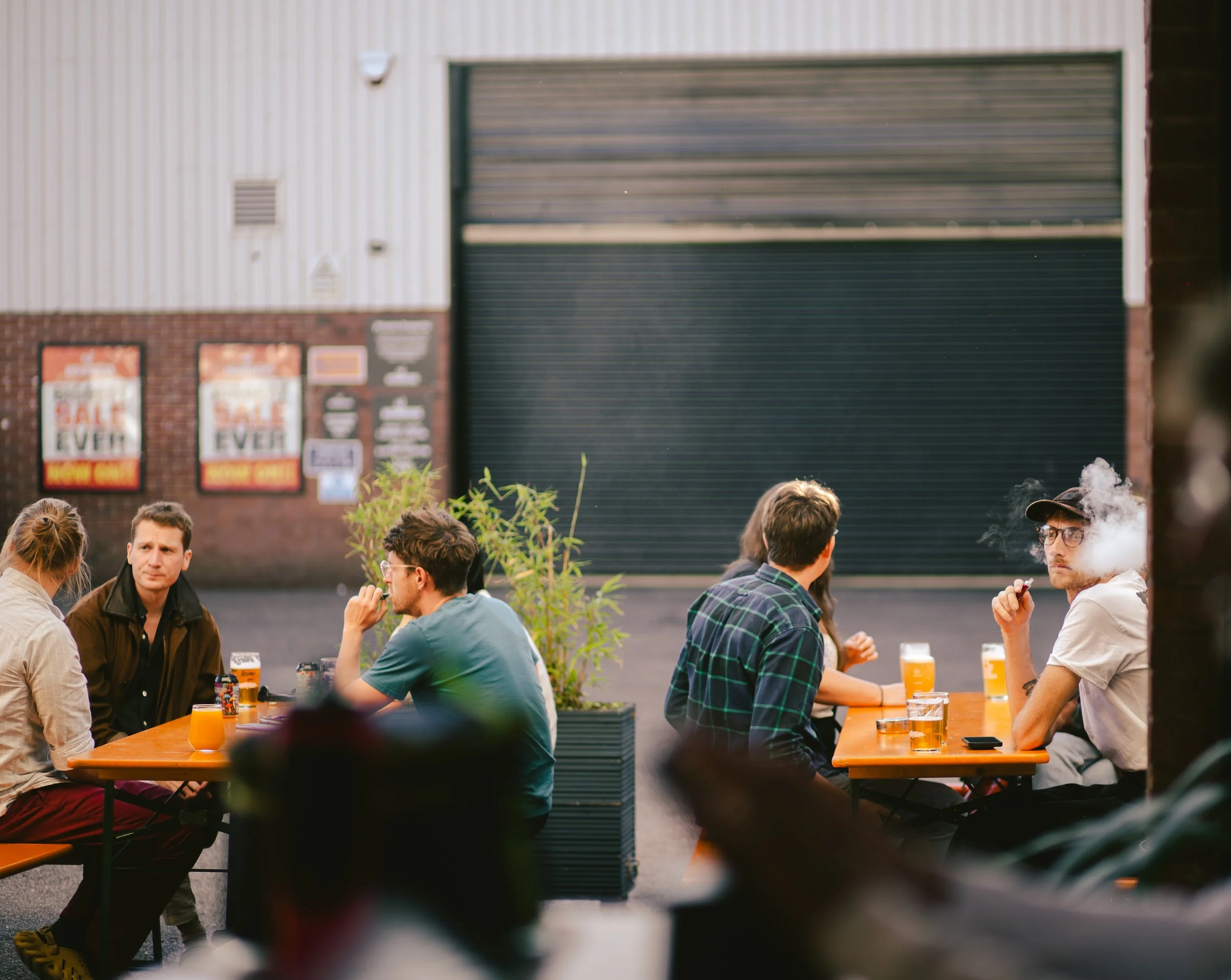 People sitting at outdoor tables drinking beer and smoking, with a closed garage door in background.