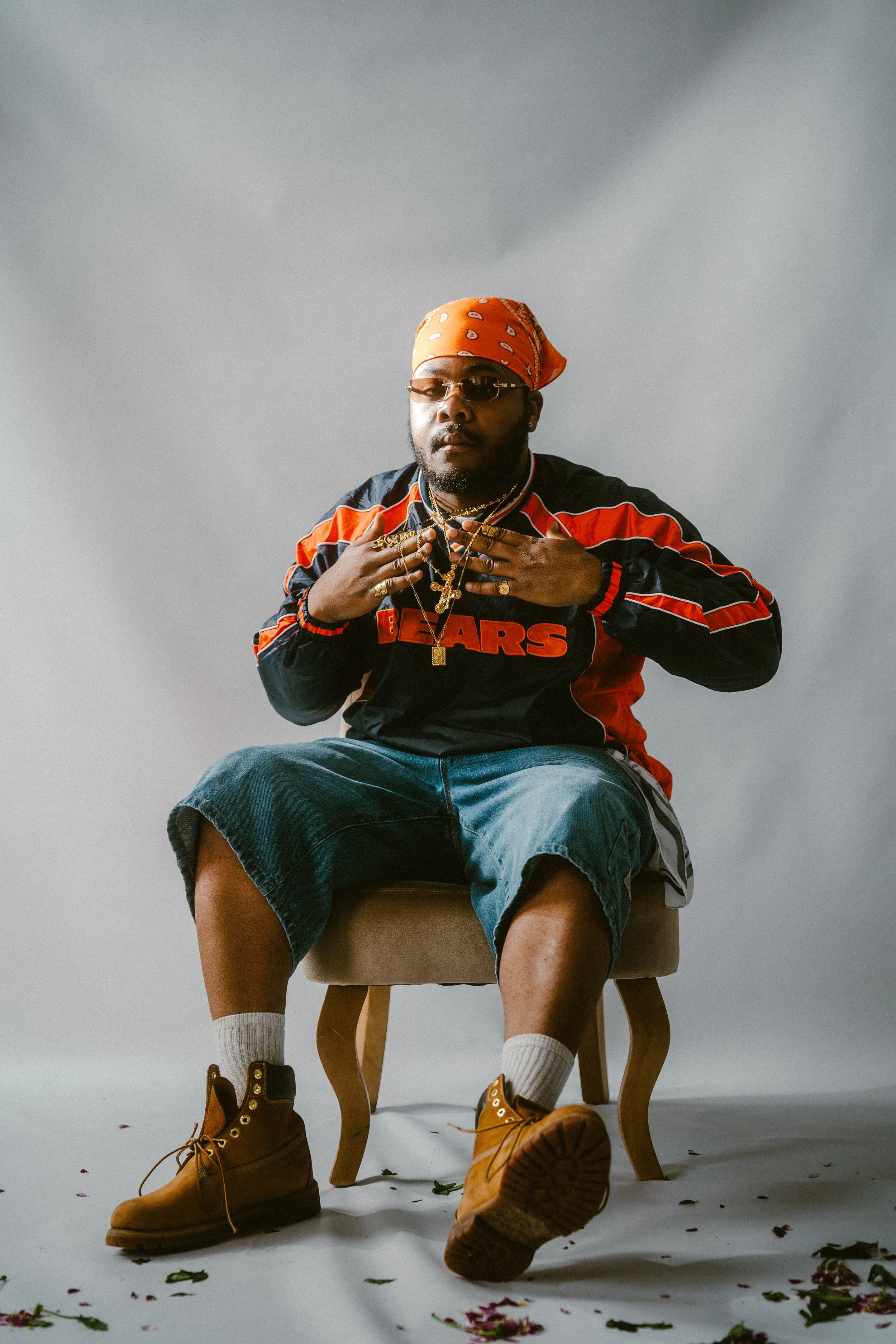 A man seated on a stool in front of a plain gray background, wearing sunglasses, an orange bandana, a black and red sports jacket, denim shorts, white socks, and tan boots, with his hands on his chest adorned with several gold rings and necklaces.