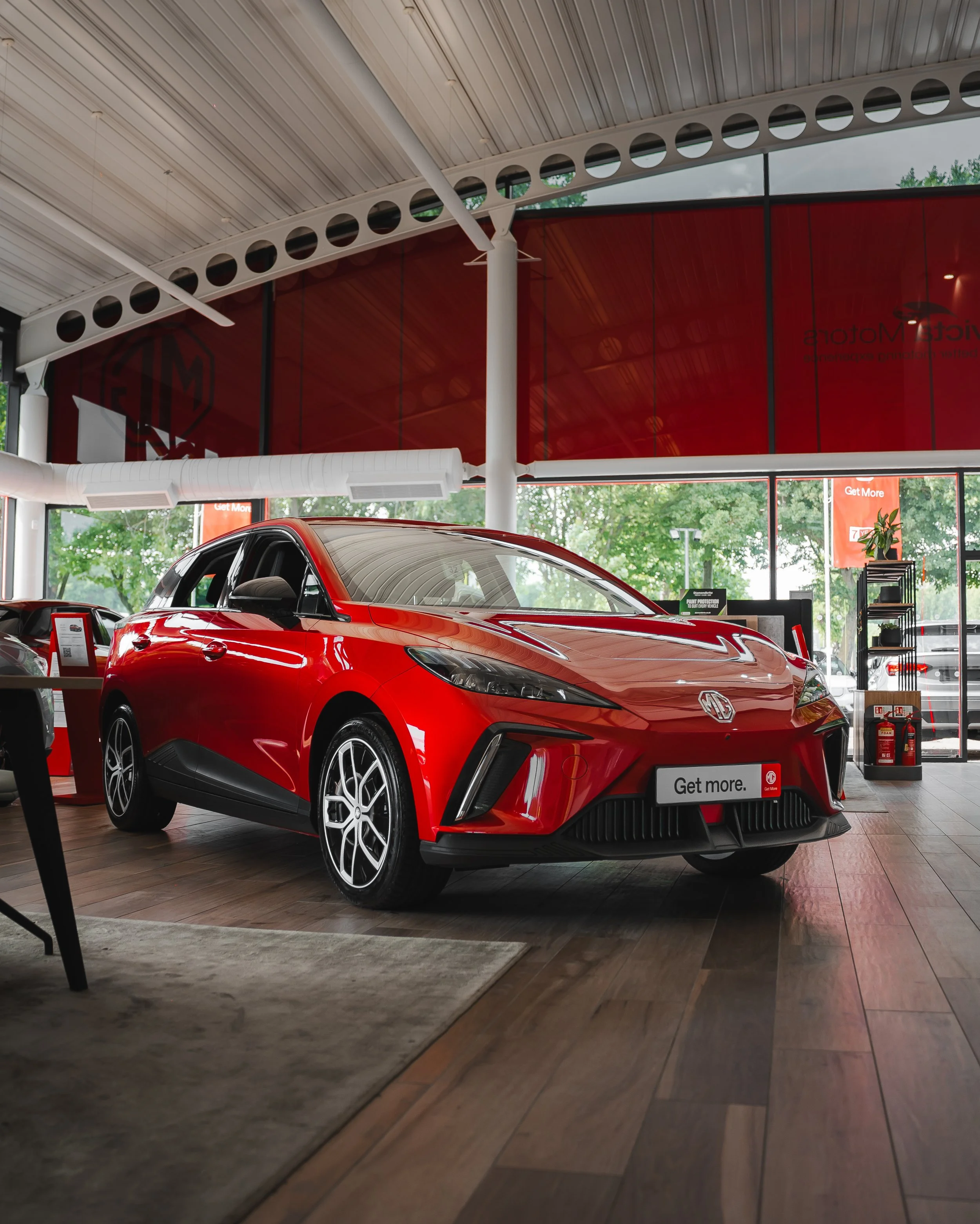 Red MG electric car inside a showroom with wooden flooring and large windows showing trees outside.