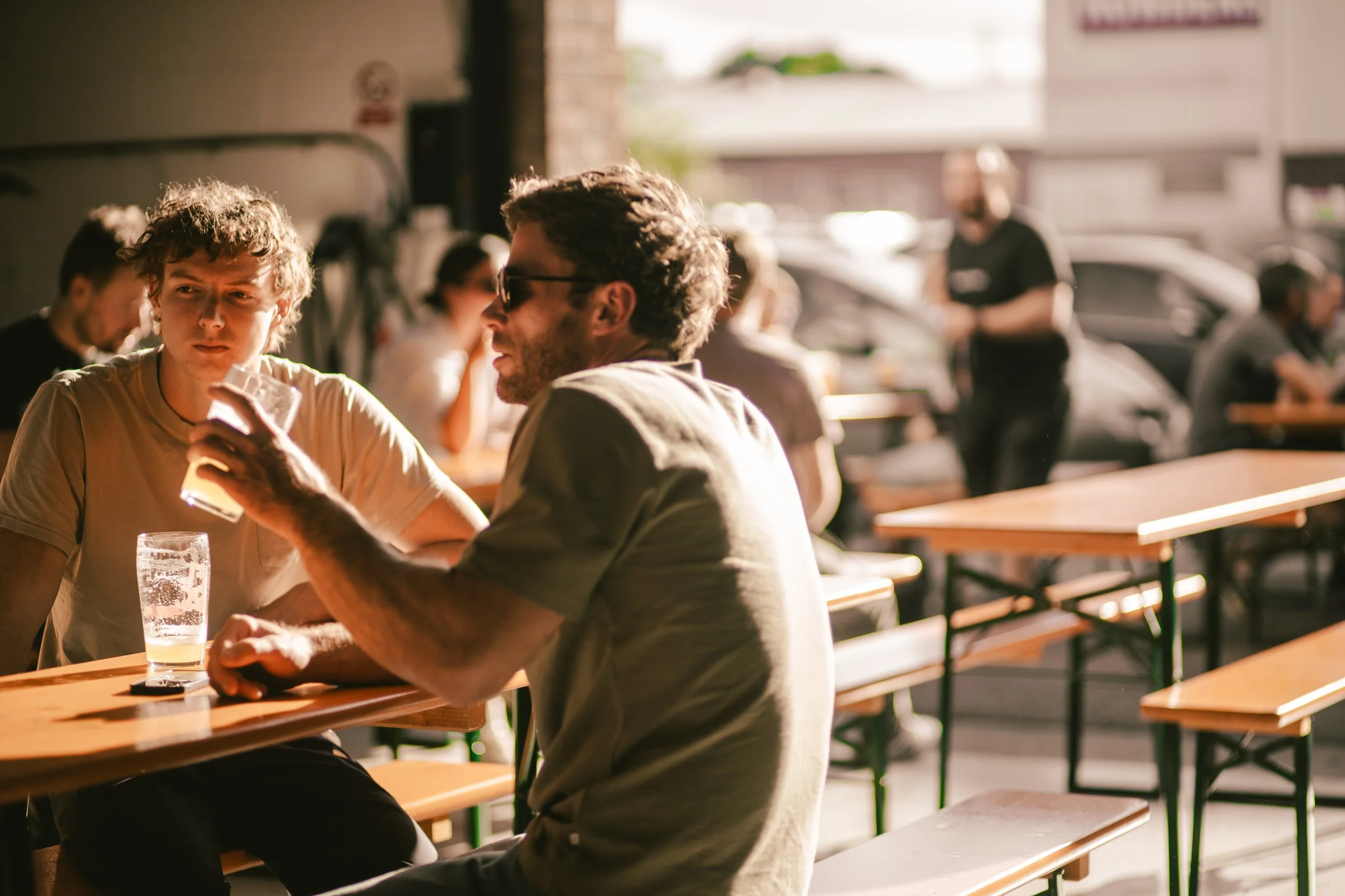 Two men sitting at an outdoor café, engaged in conversation, with drinks on the table and sunlight illuminating the scene.