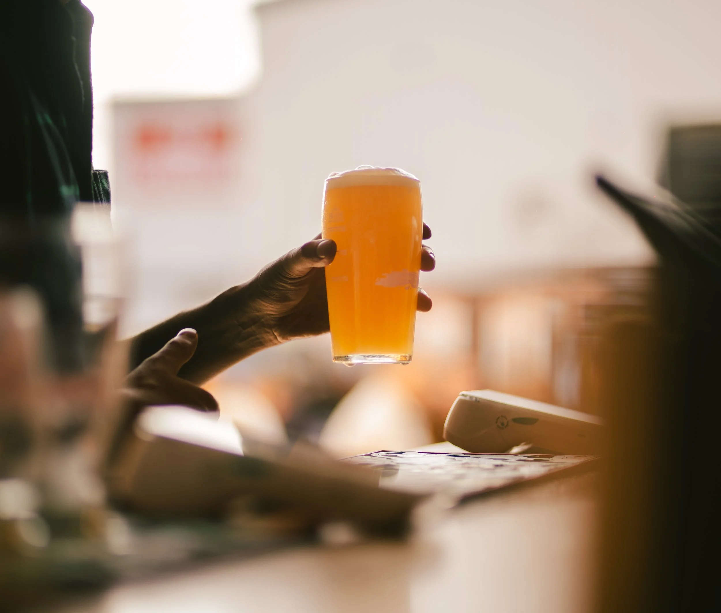 A hand holding a pint glass of foamy beer against a bright, blurred background.