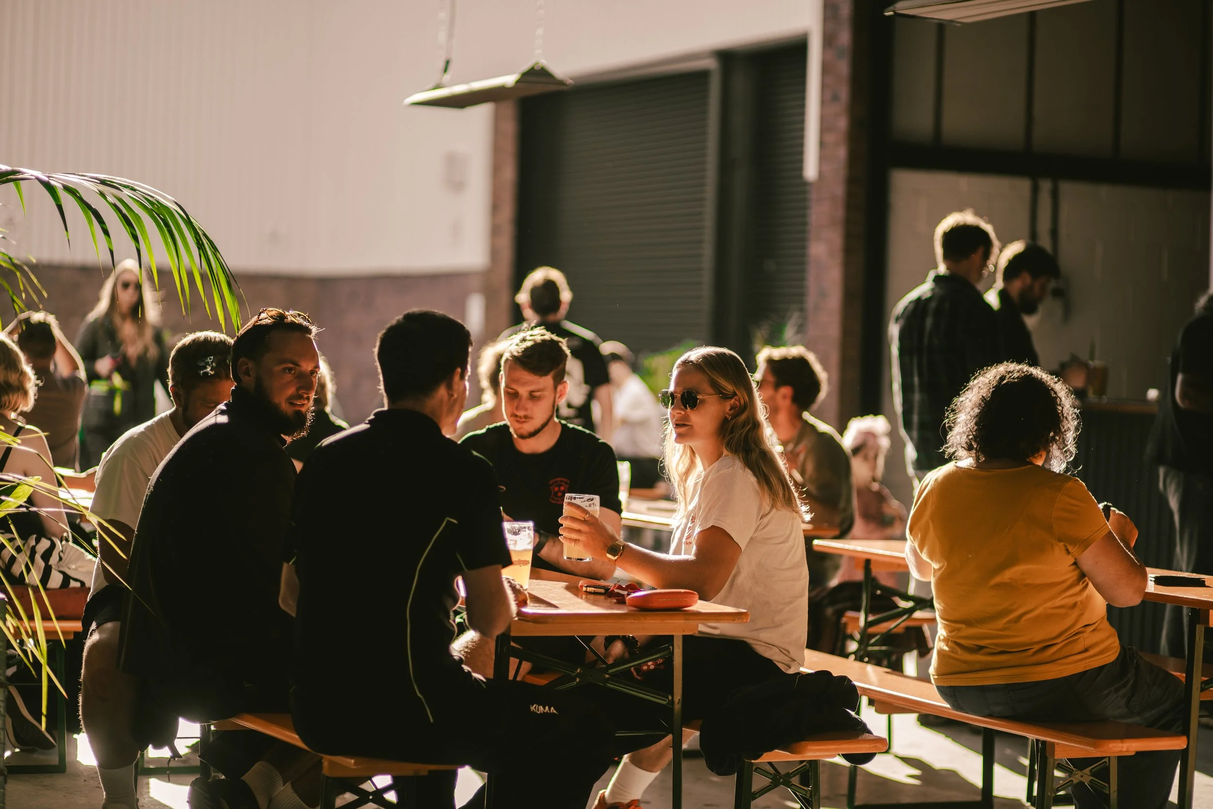 People sitting at outdoor tables, enjoying drinks, during daylight, in a casual setting with sunlight streaming in.