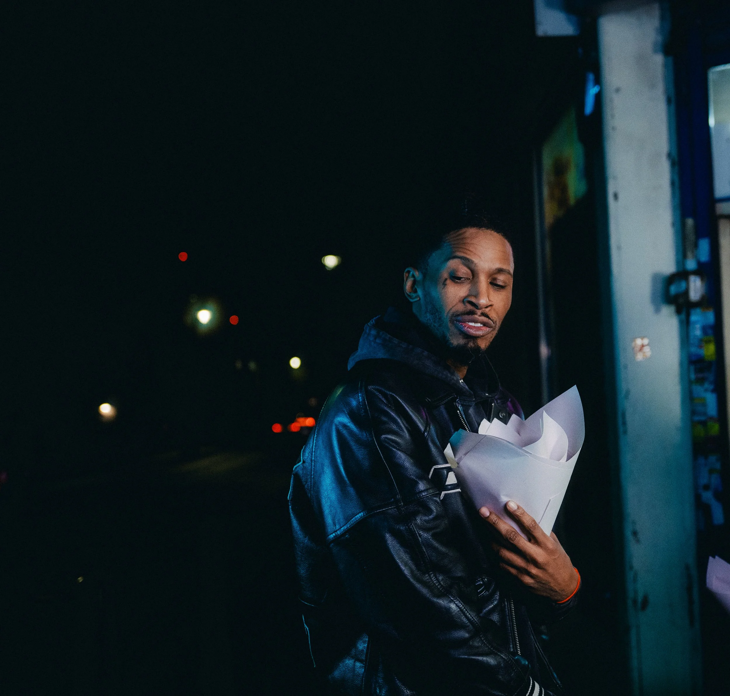 A man with a shaved head and goatee standing on a dark street at night, holding a paper bag filled with food, wearing a black leather jacket, with city lights in the background.