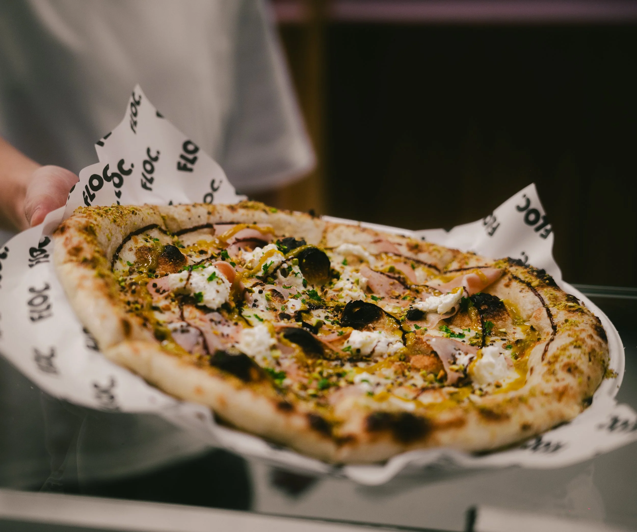 A person holding a freshly baked pizza with various toppings including cheese, olives, and ham, on branded parchment paper.