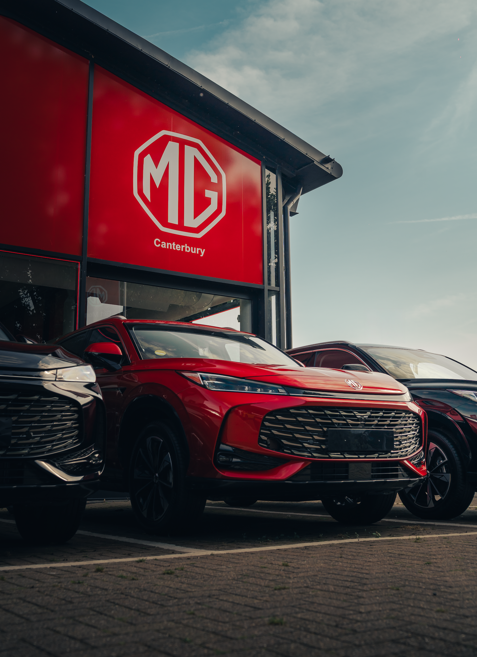 Red MG sports car parked in front of MG Canterbury dealership with two other cars, one black and one dark grey, in a row on a brick-paved lot.