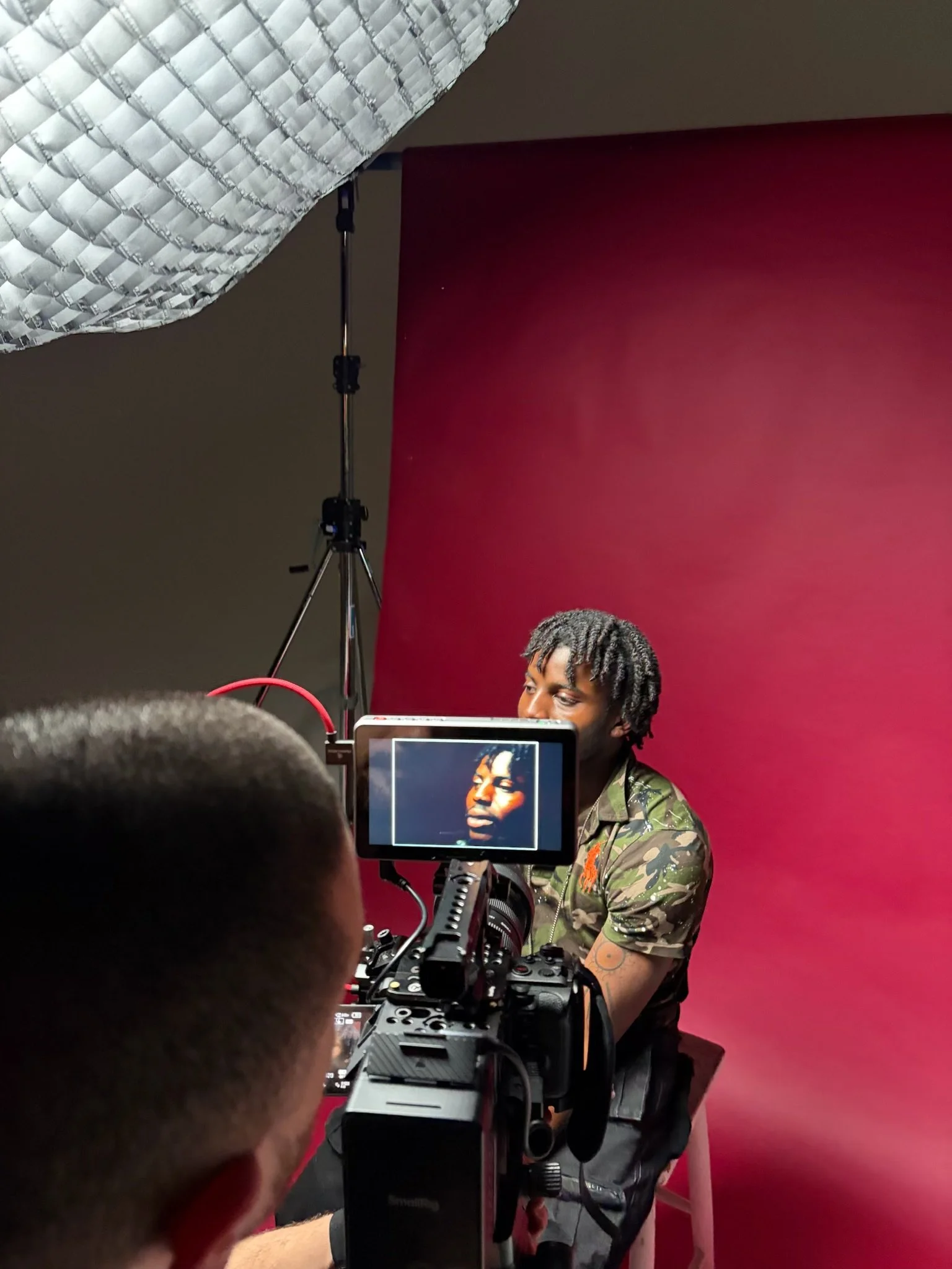 A person with a camouflage shirt sits in front of a red background while being filmed with professional camera equipment in a photography studio.