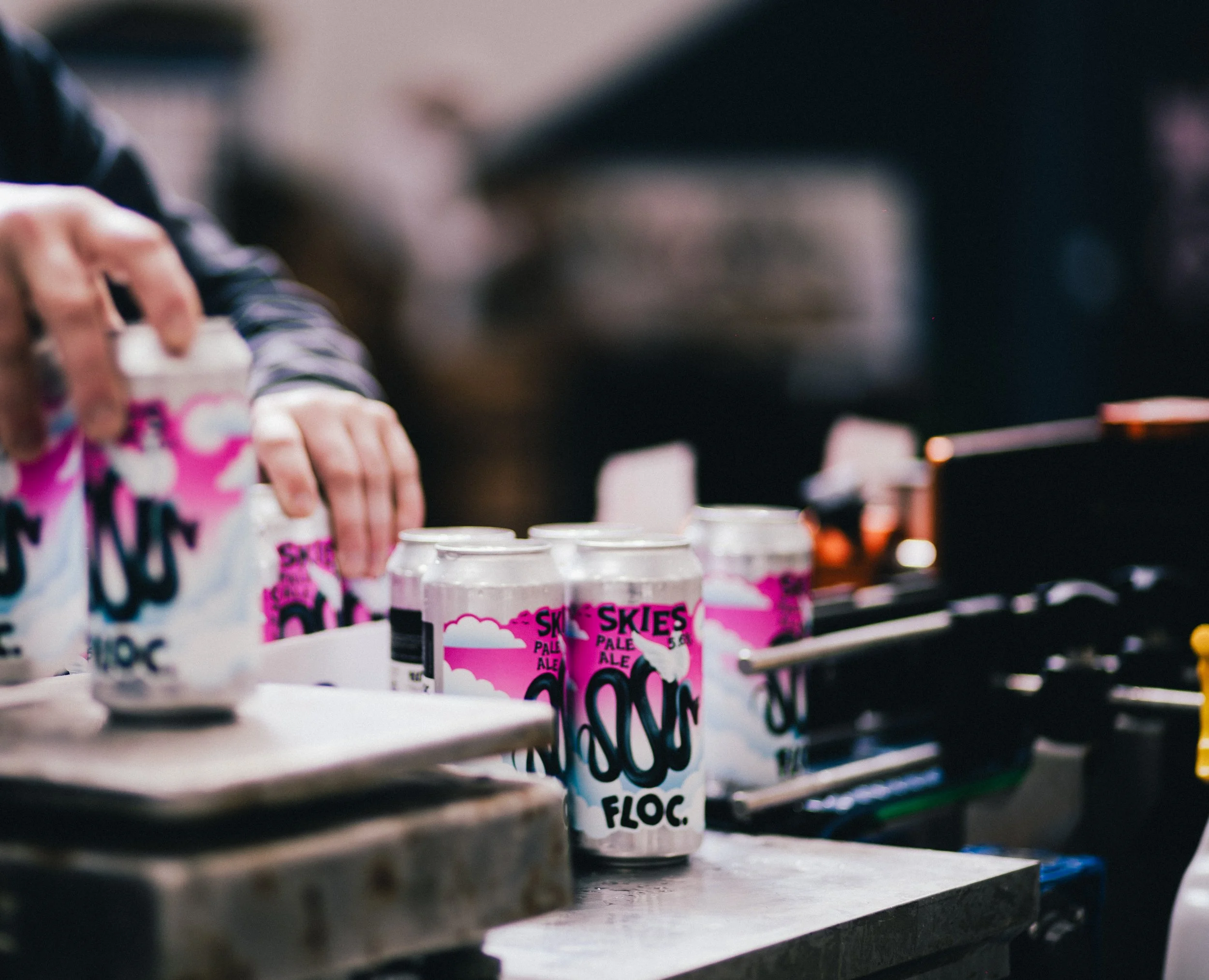 Close-up of a person hand pack cans of Skies Pale Ale with pink and white label, set on a metal surface, with a blurry background.