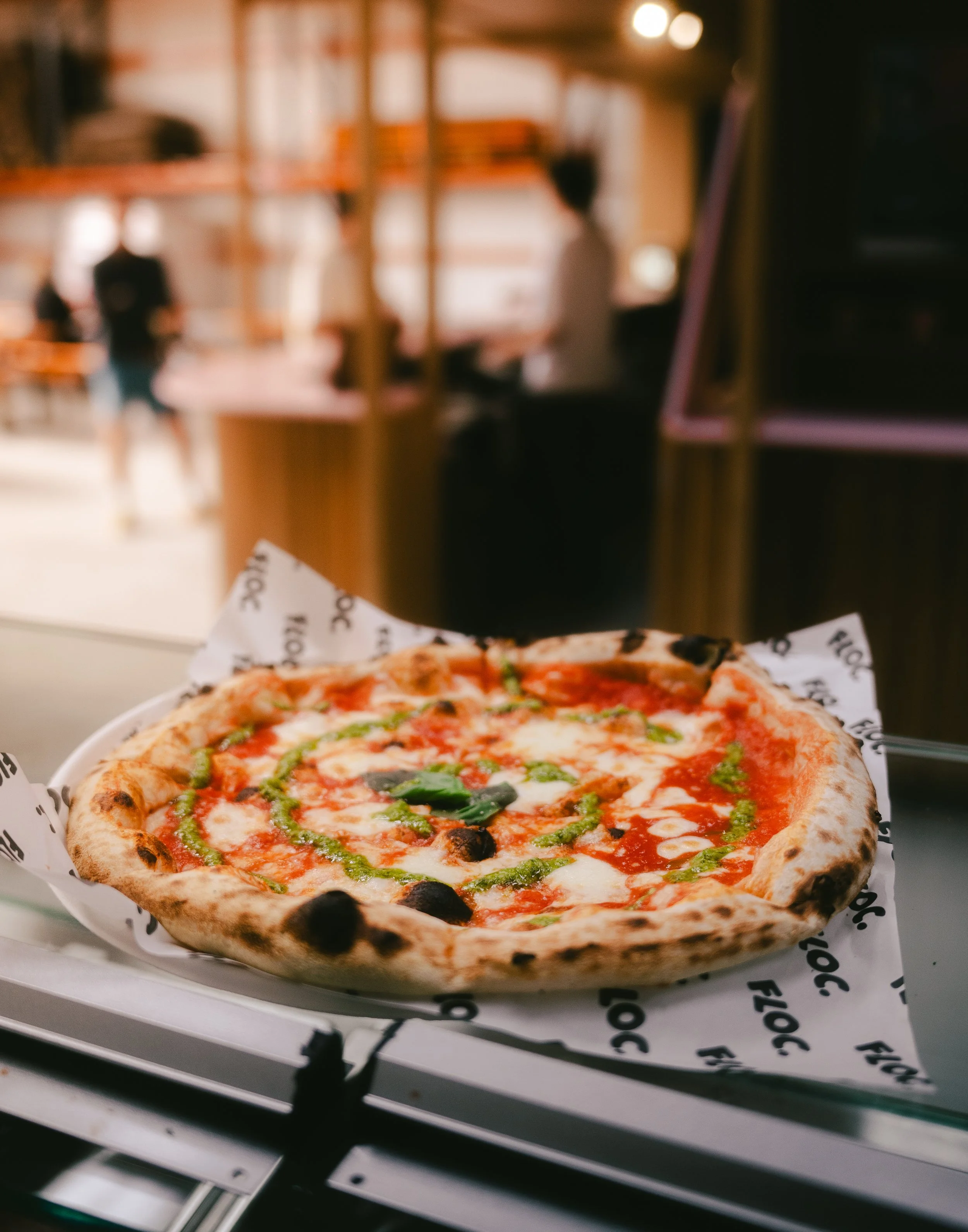 A hot pizza with cheese, tomato sauce, and green pesto, on a plate, inside a restaurant with blurred background.