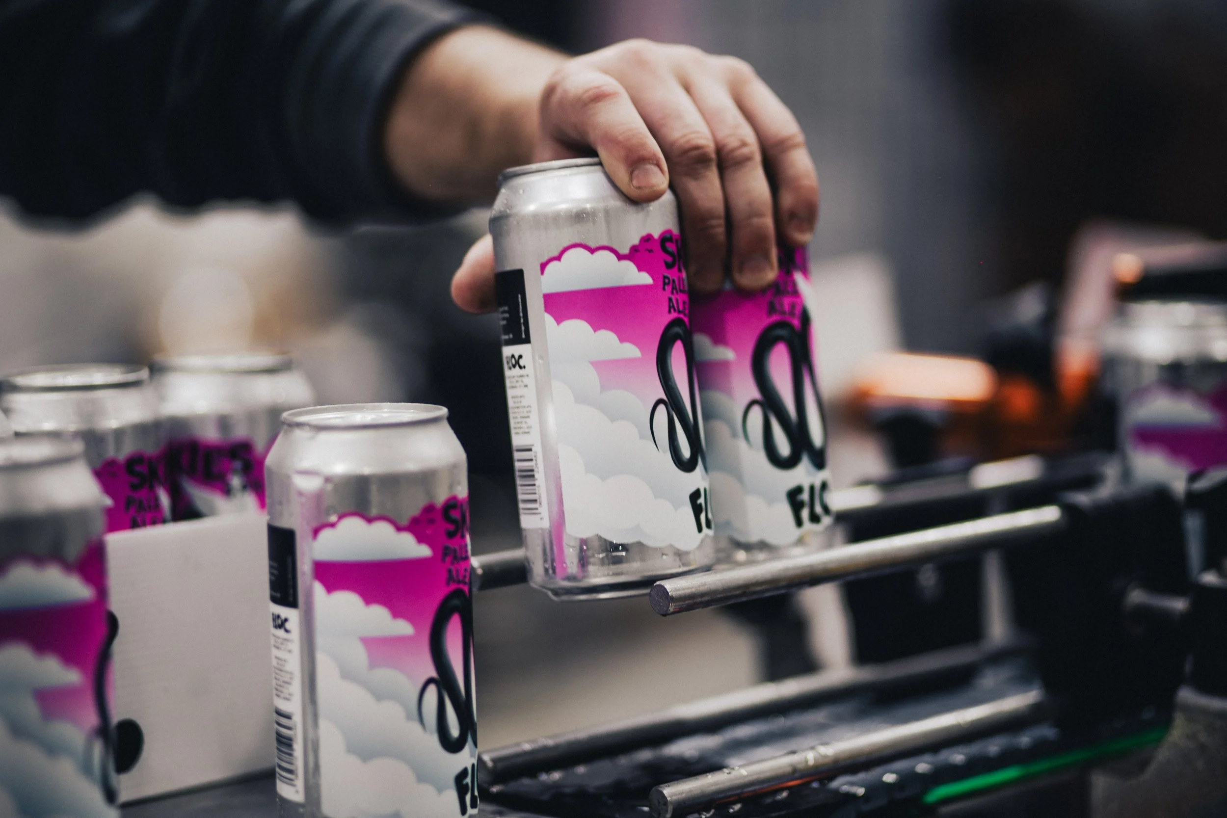 A person's hand is holding a can of White Claw flavored seltzer water, with others on a production line in the background.