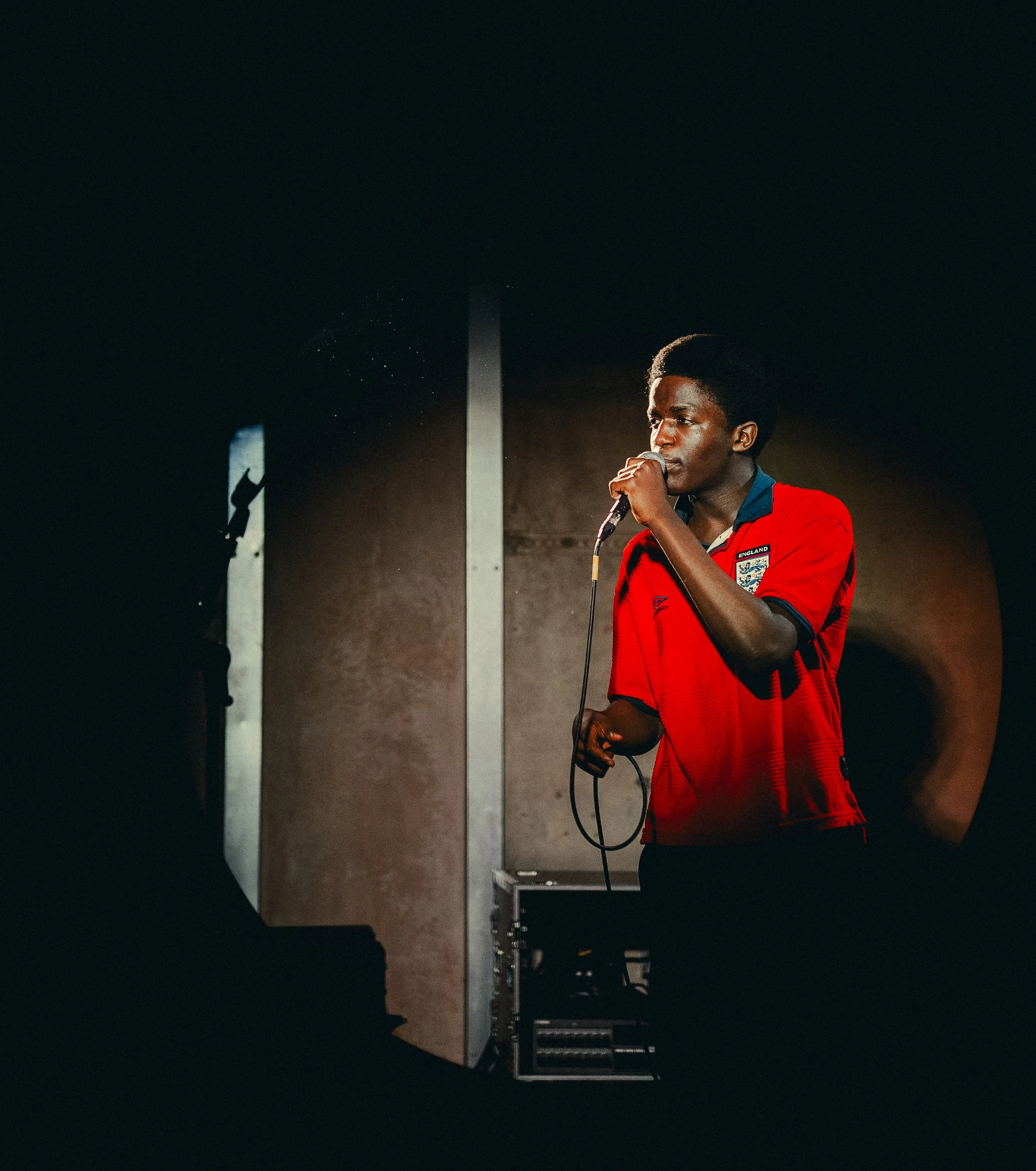 A young man with dark skin and an afro hairstyle singing into a microphone on stage, wearing a red sports jersey with an England badge.