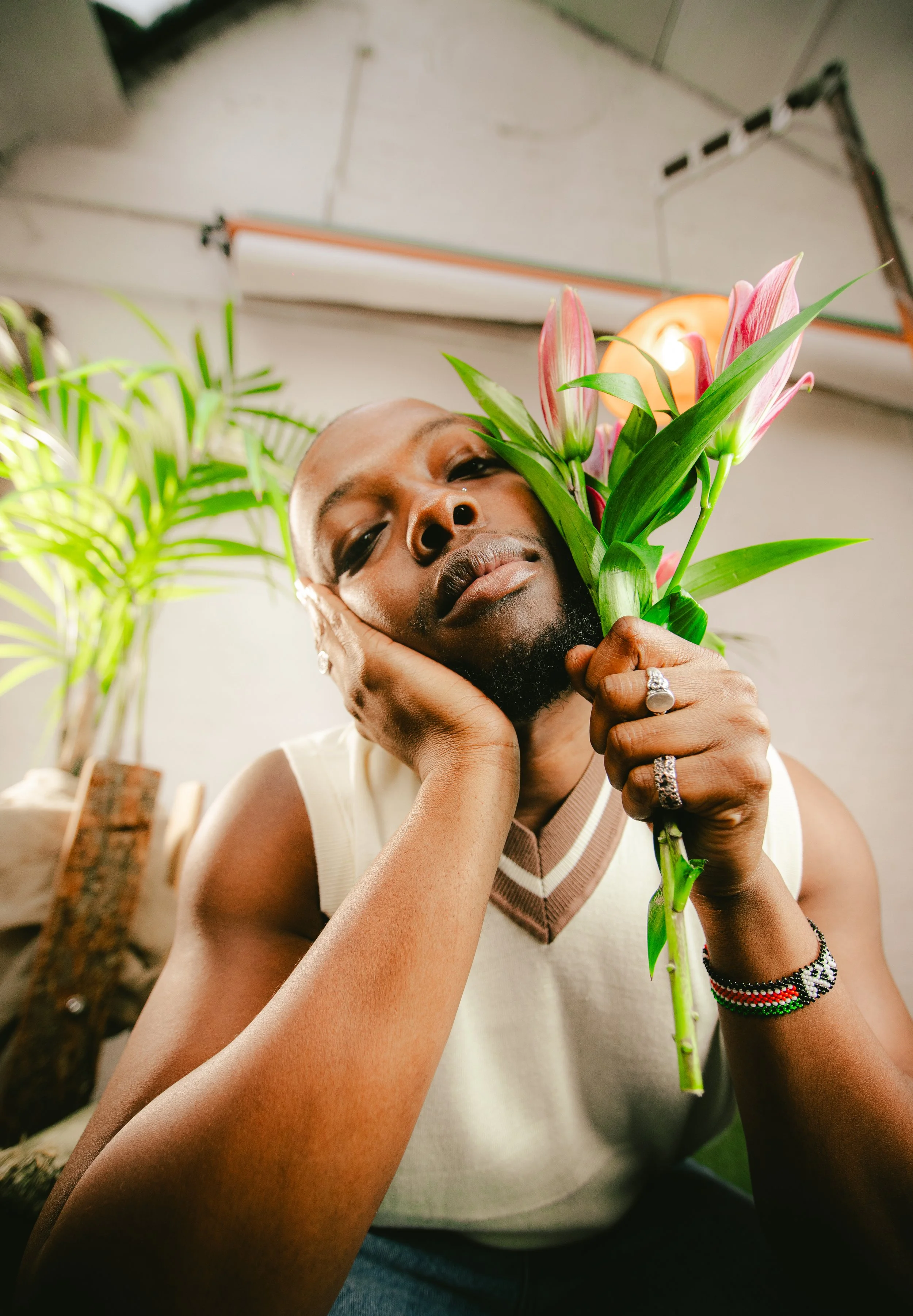 A person holding a bouquet of pink lilies, resting their chin on their hand, with a contemplative expression in an indoor setting with plants in the background.