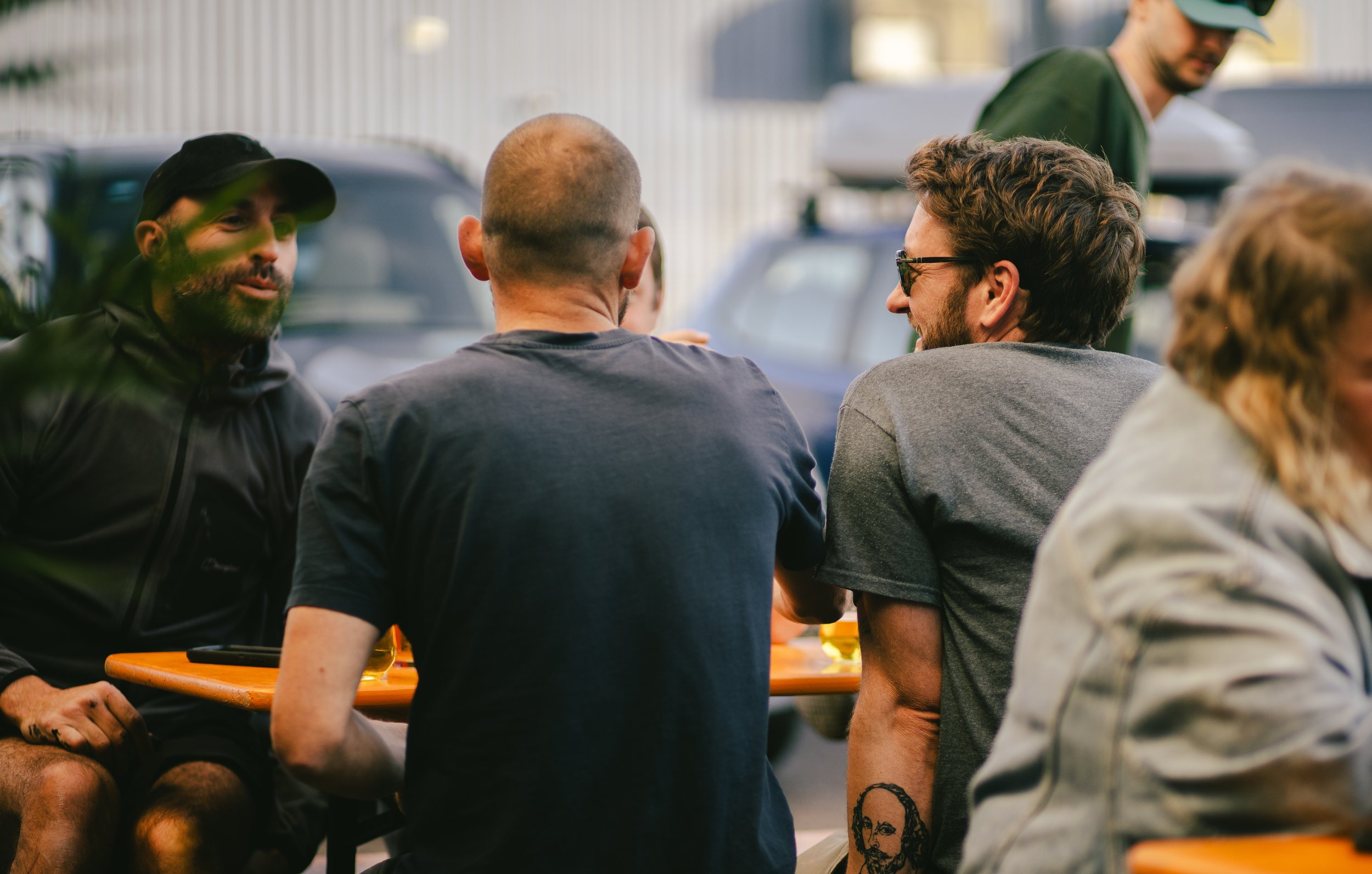 People sitting at outdoor table, engaged in conversation, in an urban setting with cars in background.