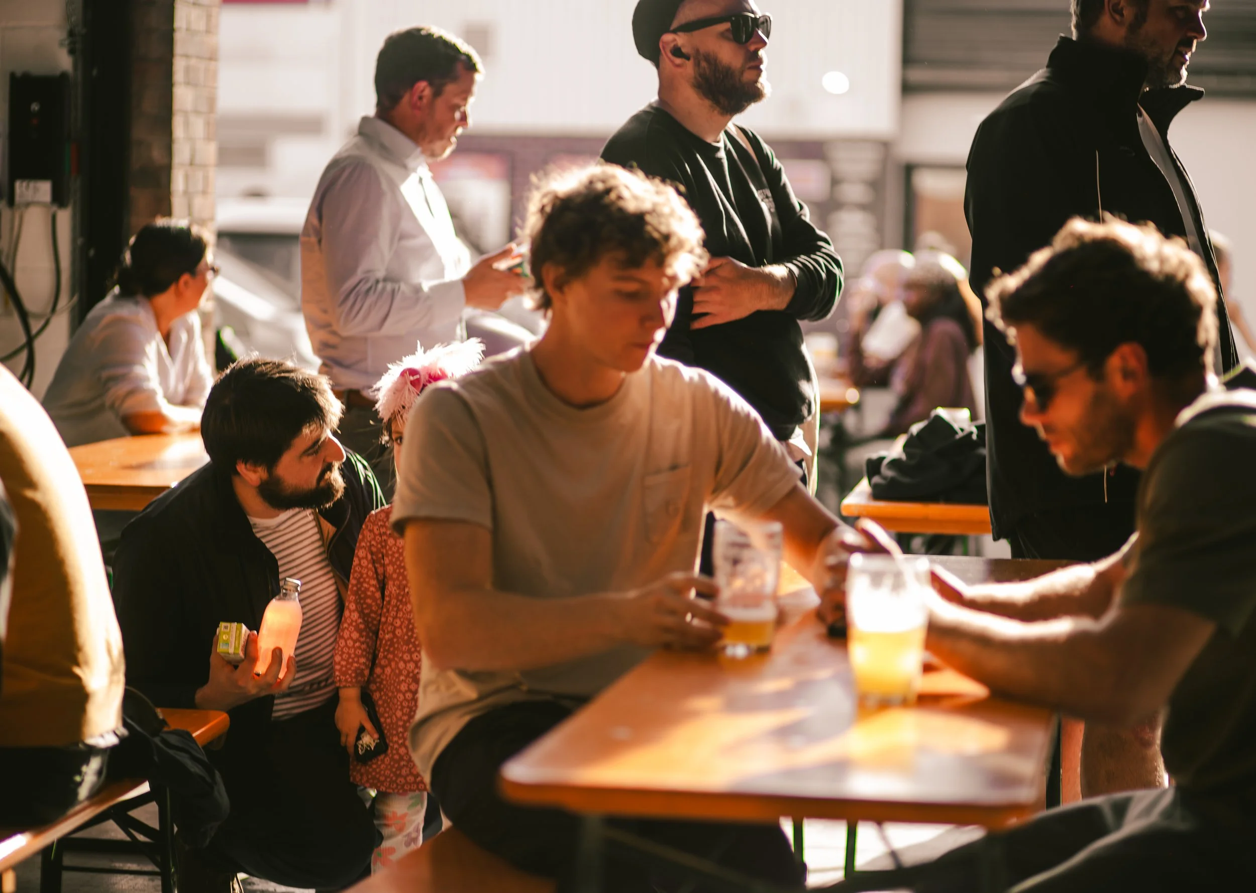 People sitting and standing inside a brightly lit cafe, some engaged with their phones and drinks, others talking, with sunlight streaming through the windows.