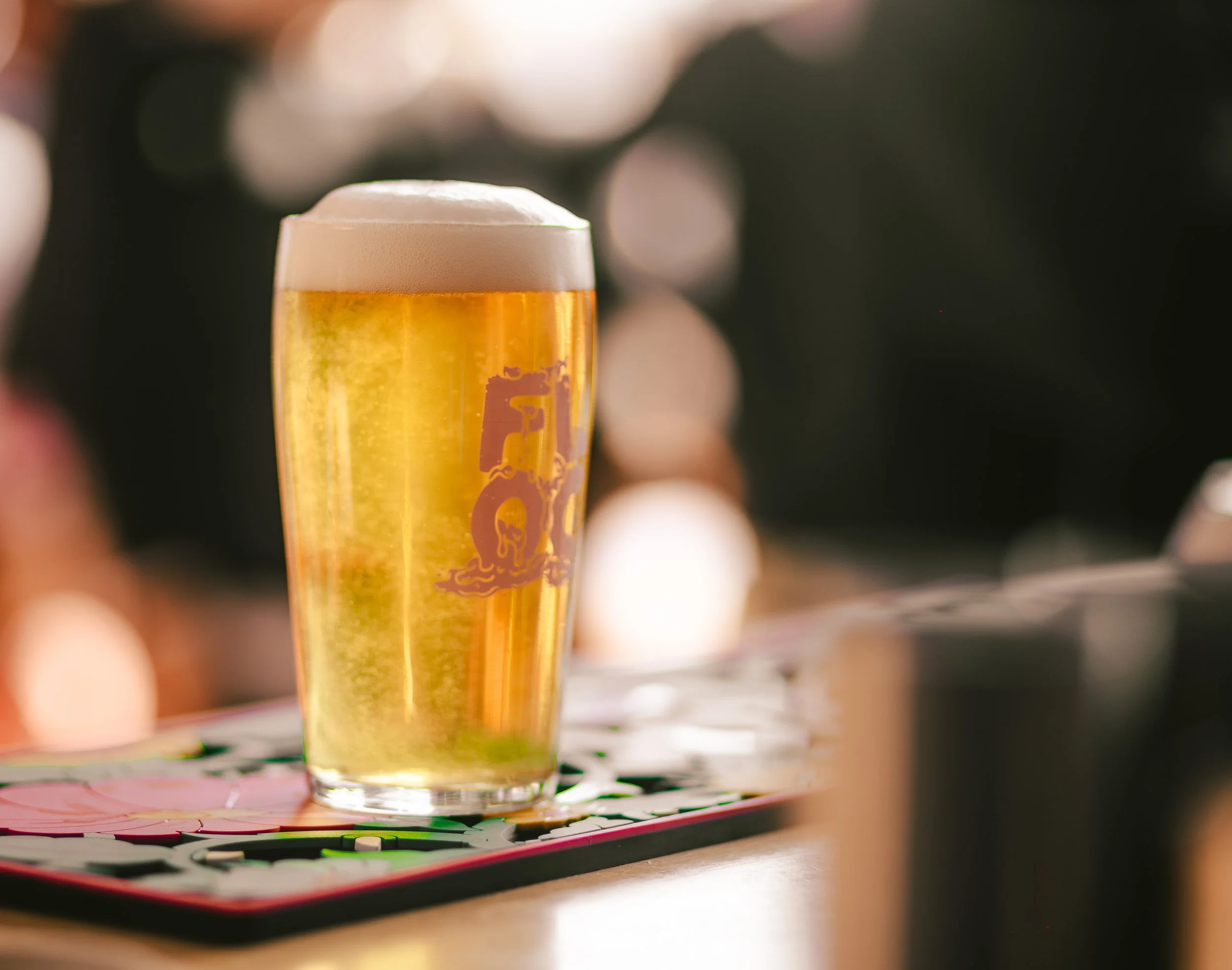 A tall glass of light-colored beer with a frothy head, placed on a bar counter with a colorful mat underneath. Bokeh background.