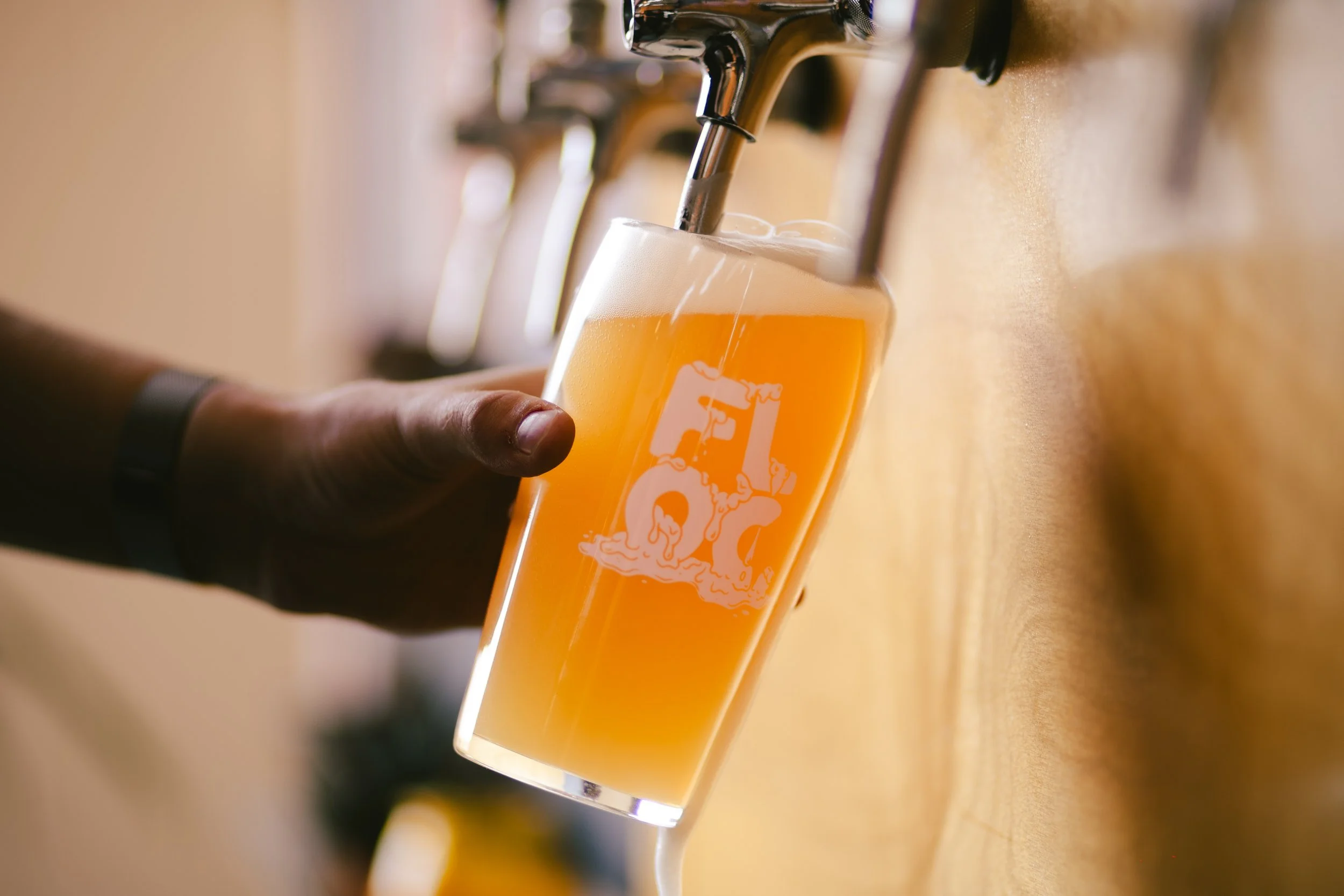 A person pours beer from a tap into a clear glass with a logo on it.