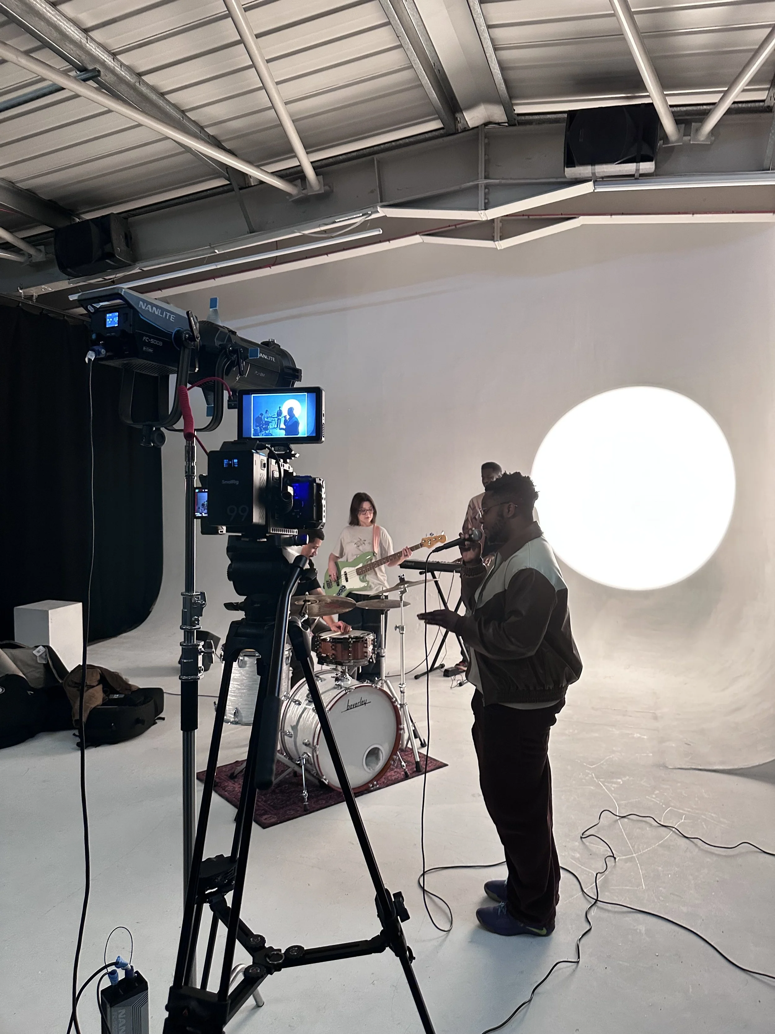 A band performing in a studio with a white backdrop and a large circular light, with a camera on a tripod recording them.