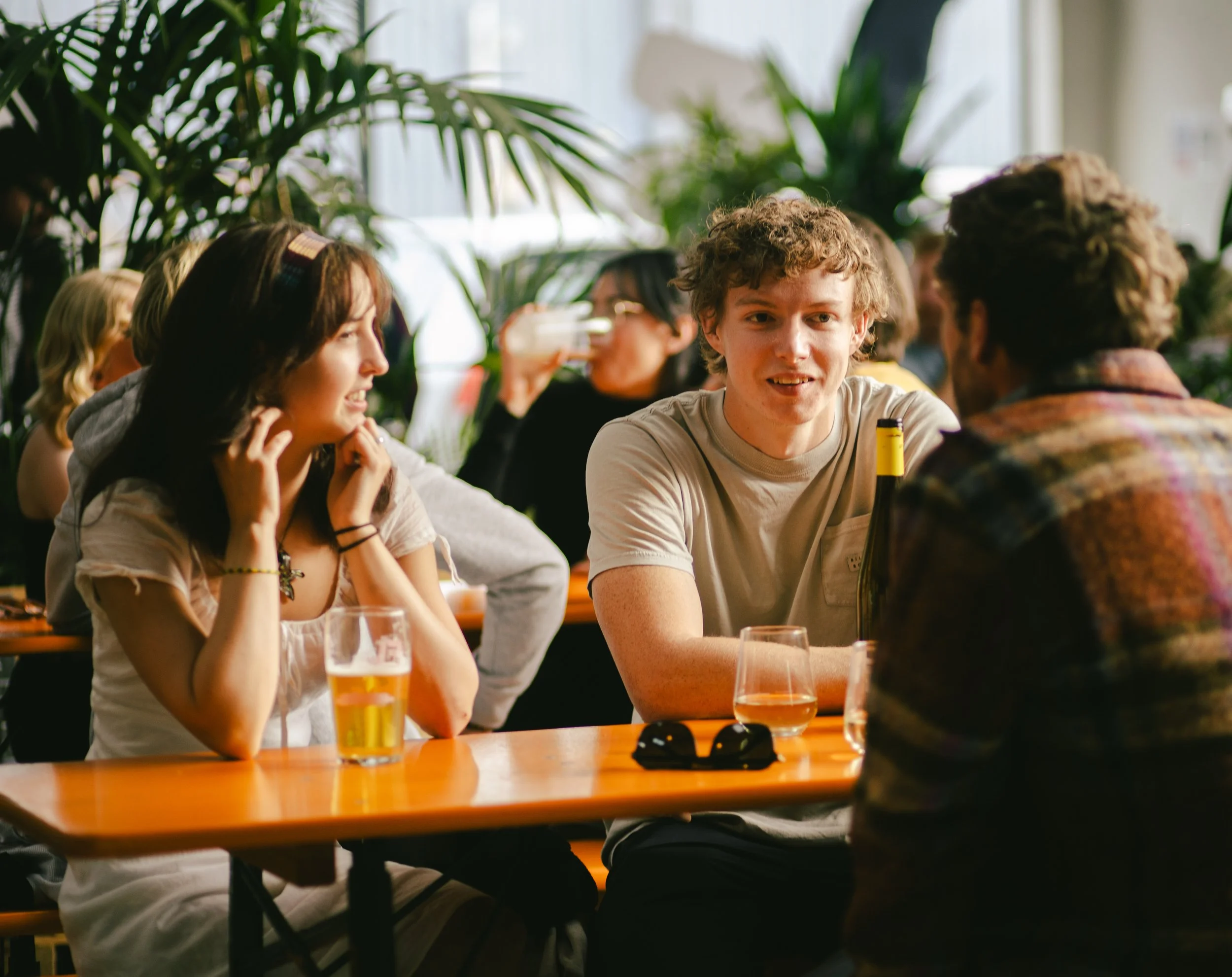 Group of young people sitting at a table in a lively, green-filled bar or restaurant, engaging in conversation and enjoying drinks.