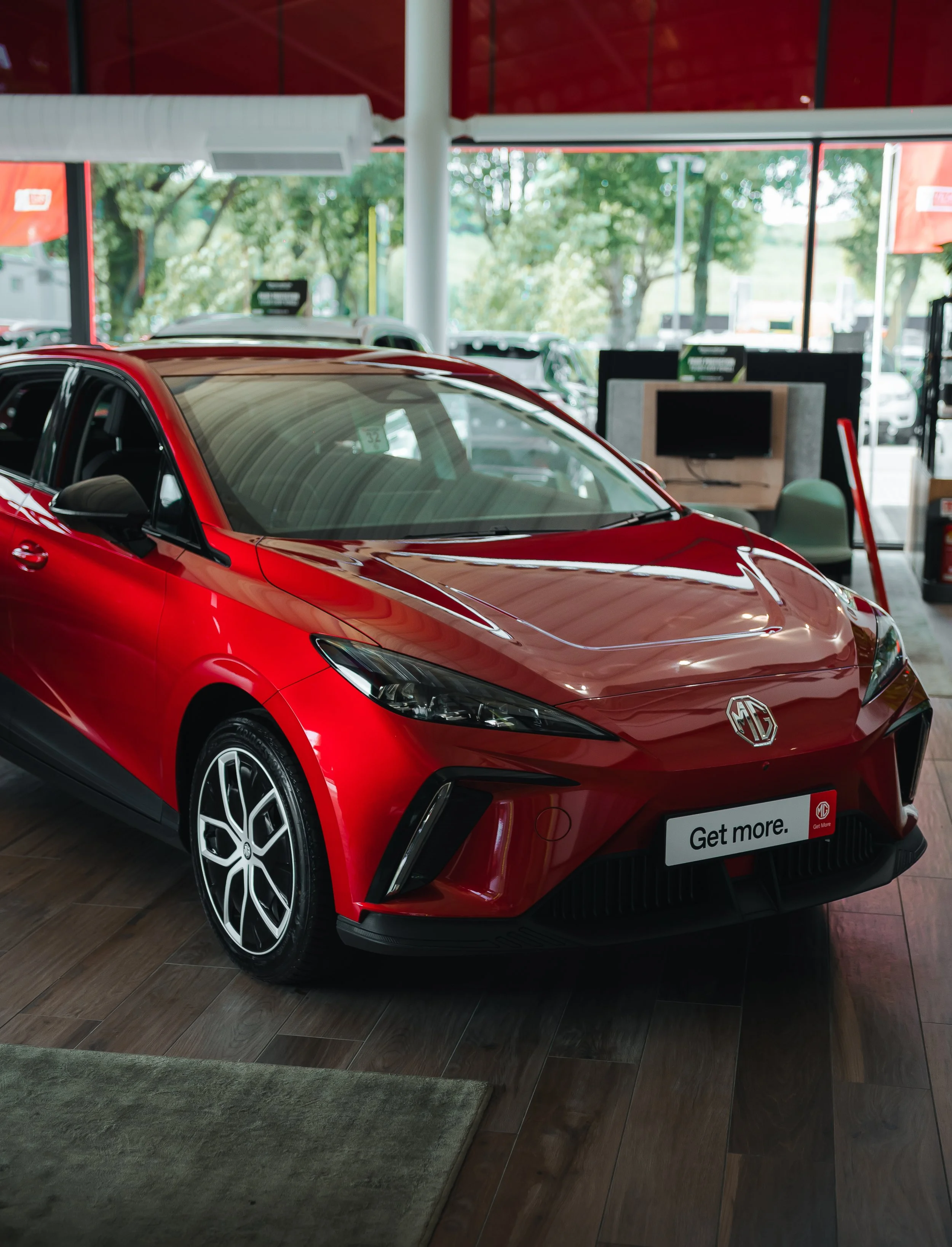 Red MG sedan on display inside a showroom with large windows showing trees outside.
